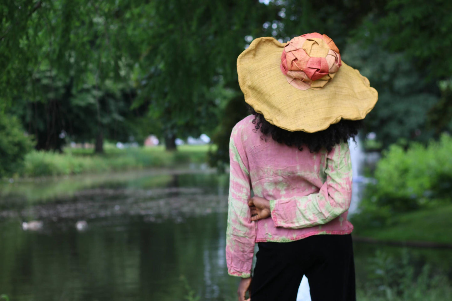 Female model wearing a vintage 90s pink and green tie-dye printe top and large straw hat, looking away from the camera by a pond.