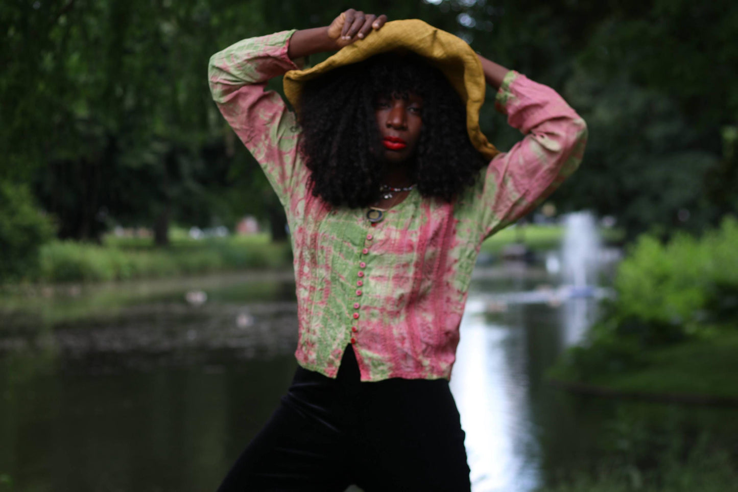 Female Model with curly black hair wearing a boho chic tie dye long sleeves blouse in pink and green and large straw hat, arms raised, posing by a pond.