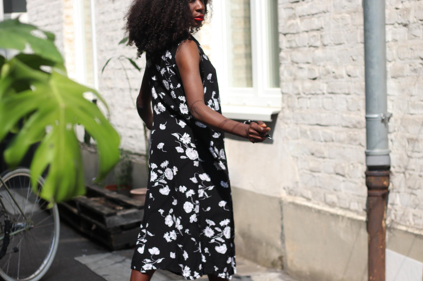 A woman with dark curly hair wearing a vintage 90s black sleeveless dress with white floral patterns stands outdoors, her back slightly turned to the camera, showcasing the side profile of the grunge inspired dress.