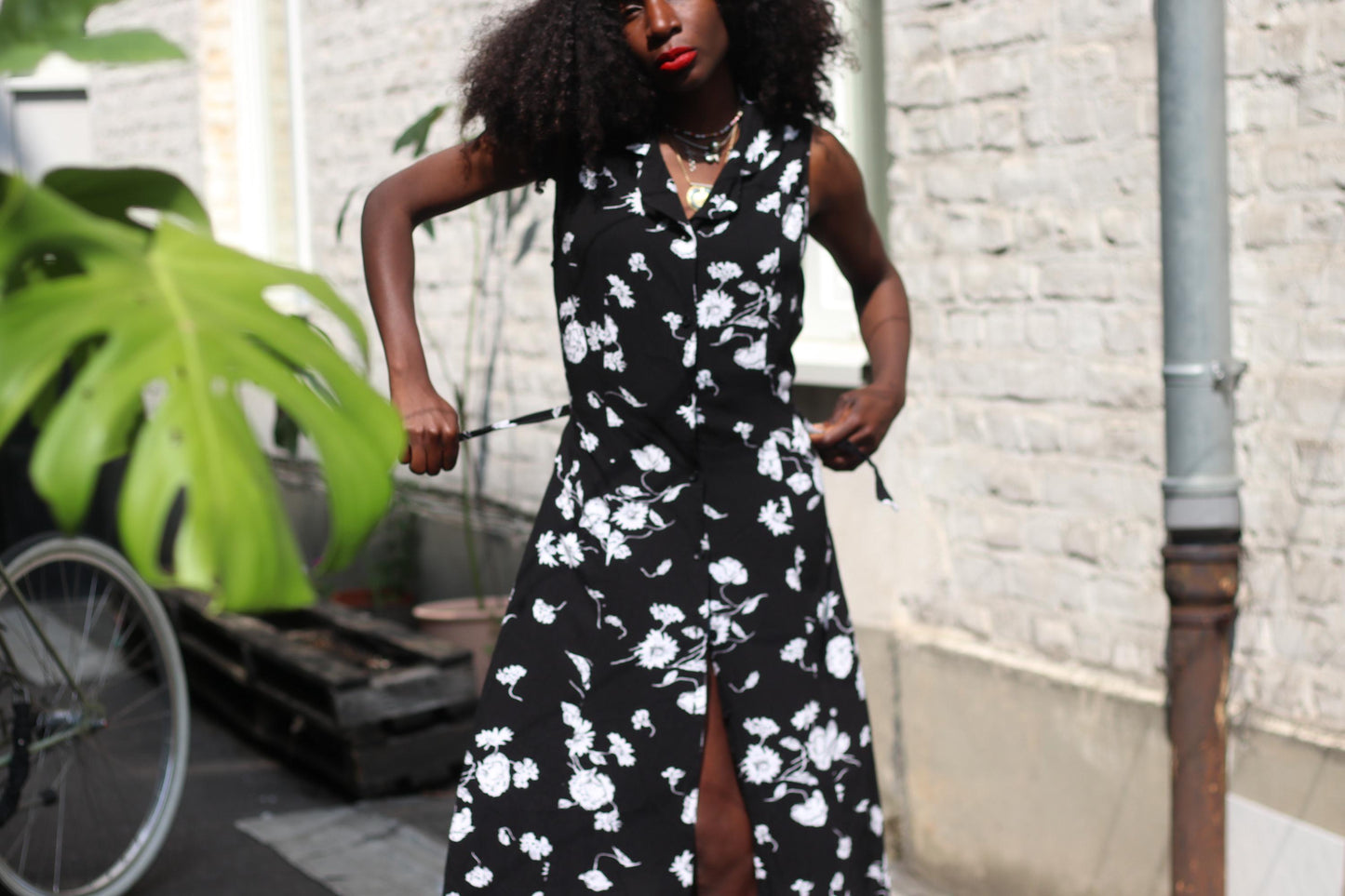 A female model.with curly black hair in a vintage black sleeveless dress from the 90s with white floral patterns stands outdoors. She showcases the dress's silhouette by holding its back tie , one in each hand.