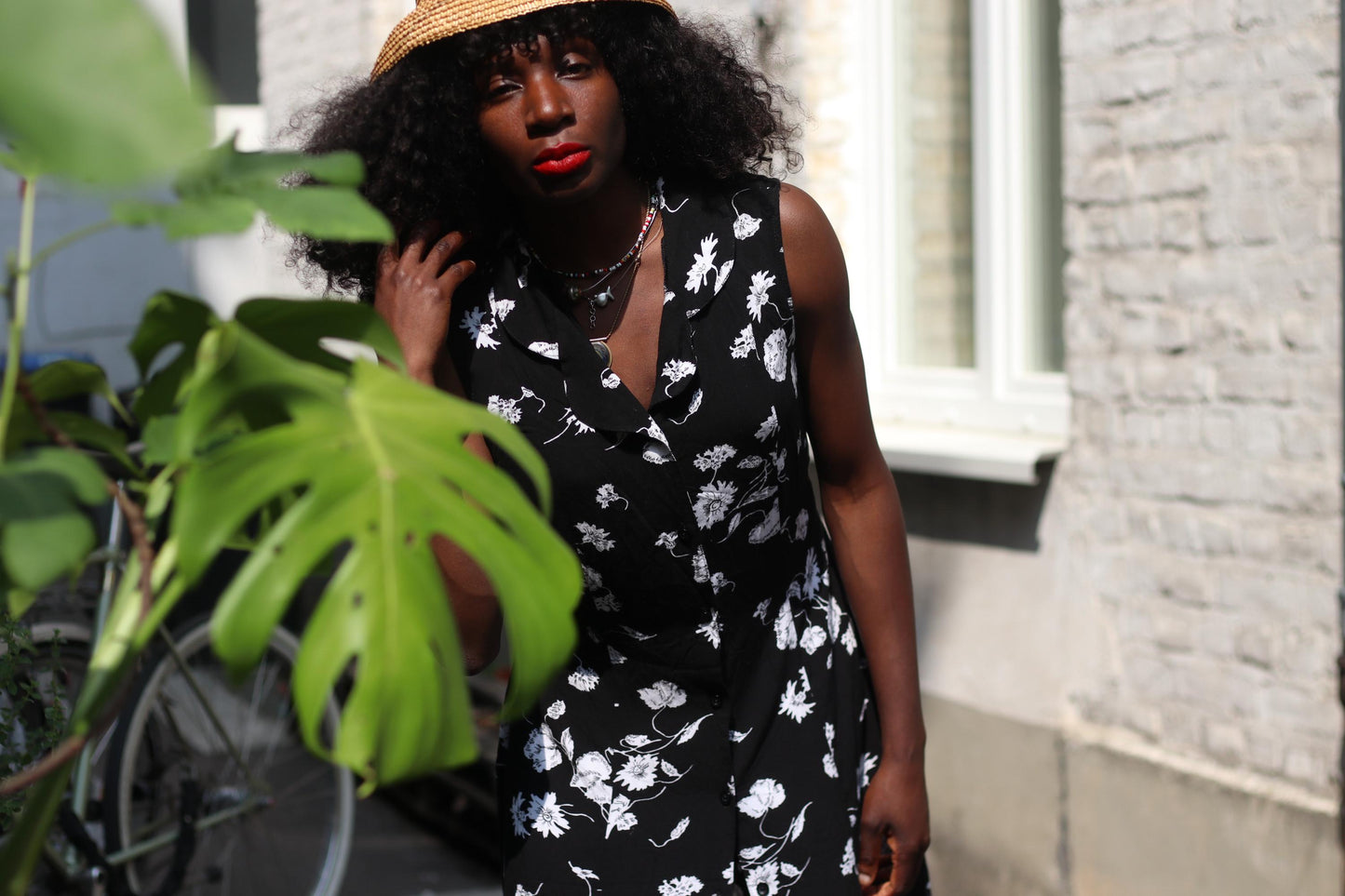 A woman wearing a black sleeveless dress with white floral patterns stands outdoors, with a green leaf from a monsera plant is in the foreground
