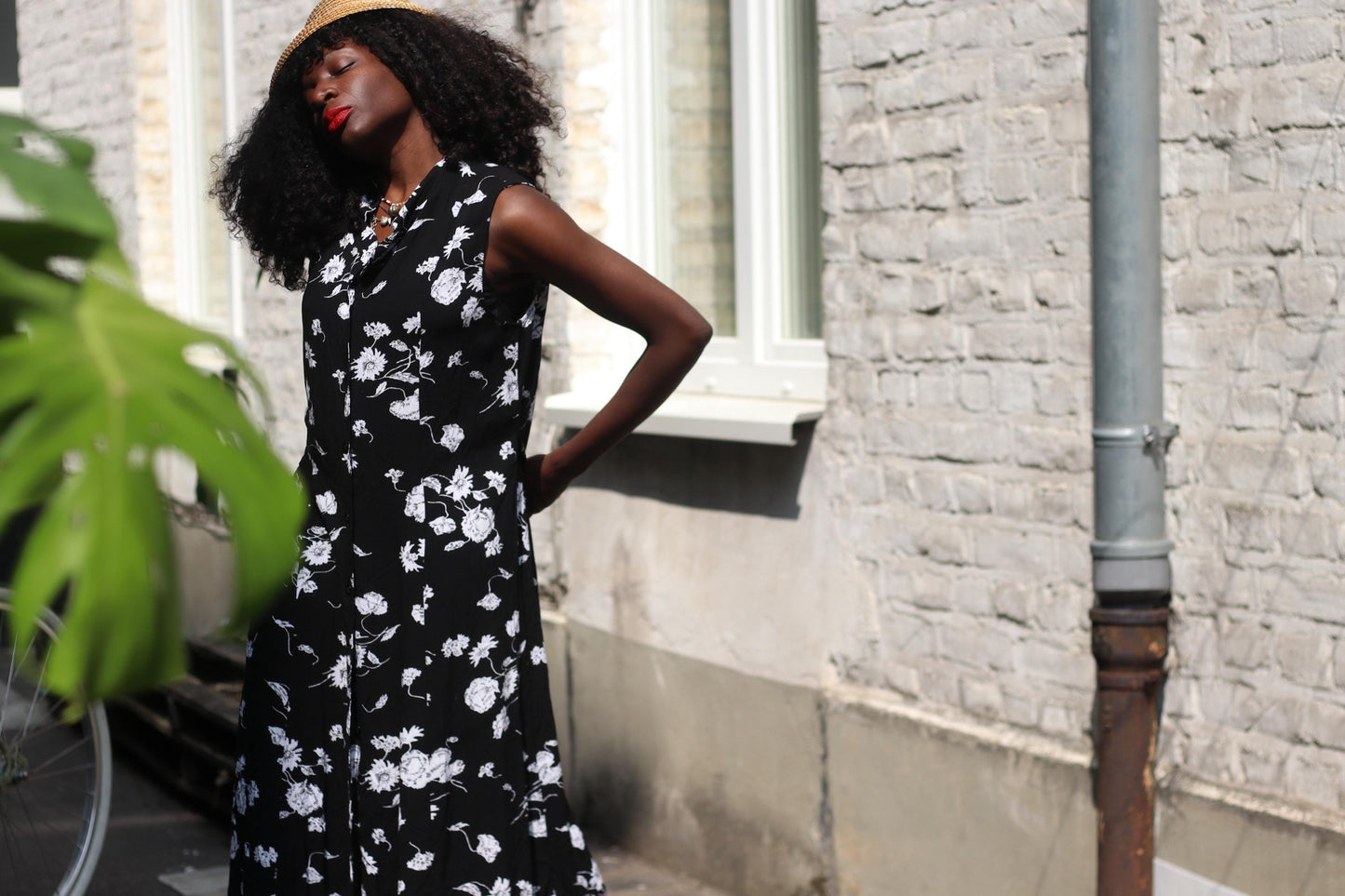 A woman wearing a vintage black sleeveless dress with white floral patterns stands outdoors, with her back slightly turned, showcasing the back of the dress.