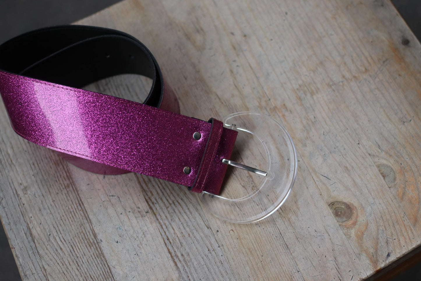 A modern-looking belt with a bold magenta glitter strap and a distinctive transparent half-moon buckle on a wide black band, laid flat on wood.