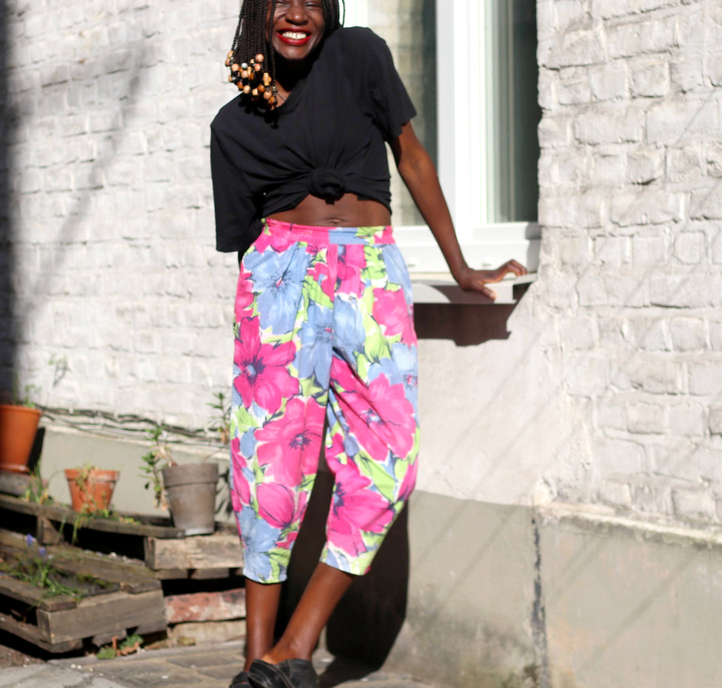 A smiling model with braids in a pair of vintage 90s tropical floral-print summer capri pants poses against a sunlit brick wall.