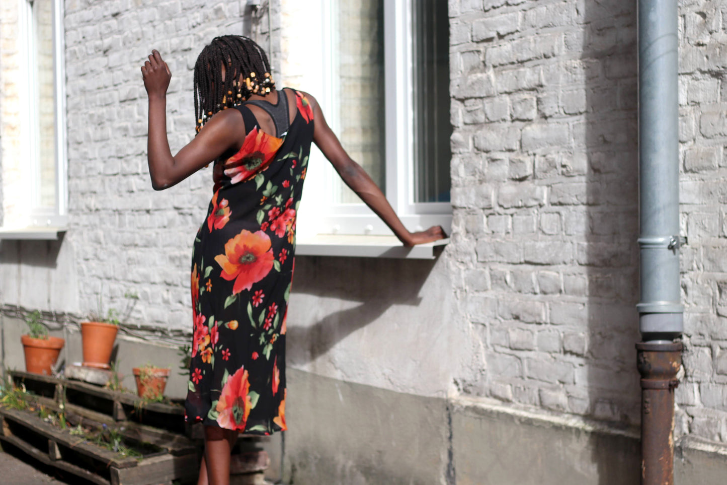 The back view of a female model wearing a 90s black semi sheer vintage sleeveless midi dress with a red and orange floral print is photographed outdoors. The background shows a white windowsill, a brick wall, terracotta pots, and a wooden pallet with plants.