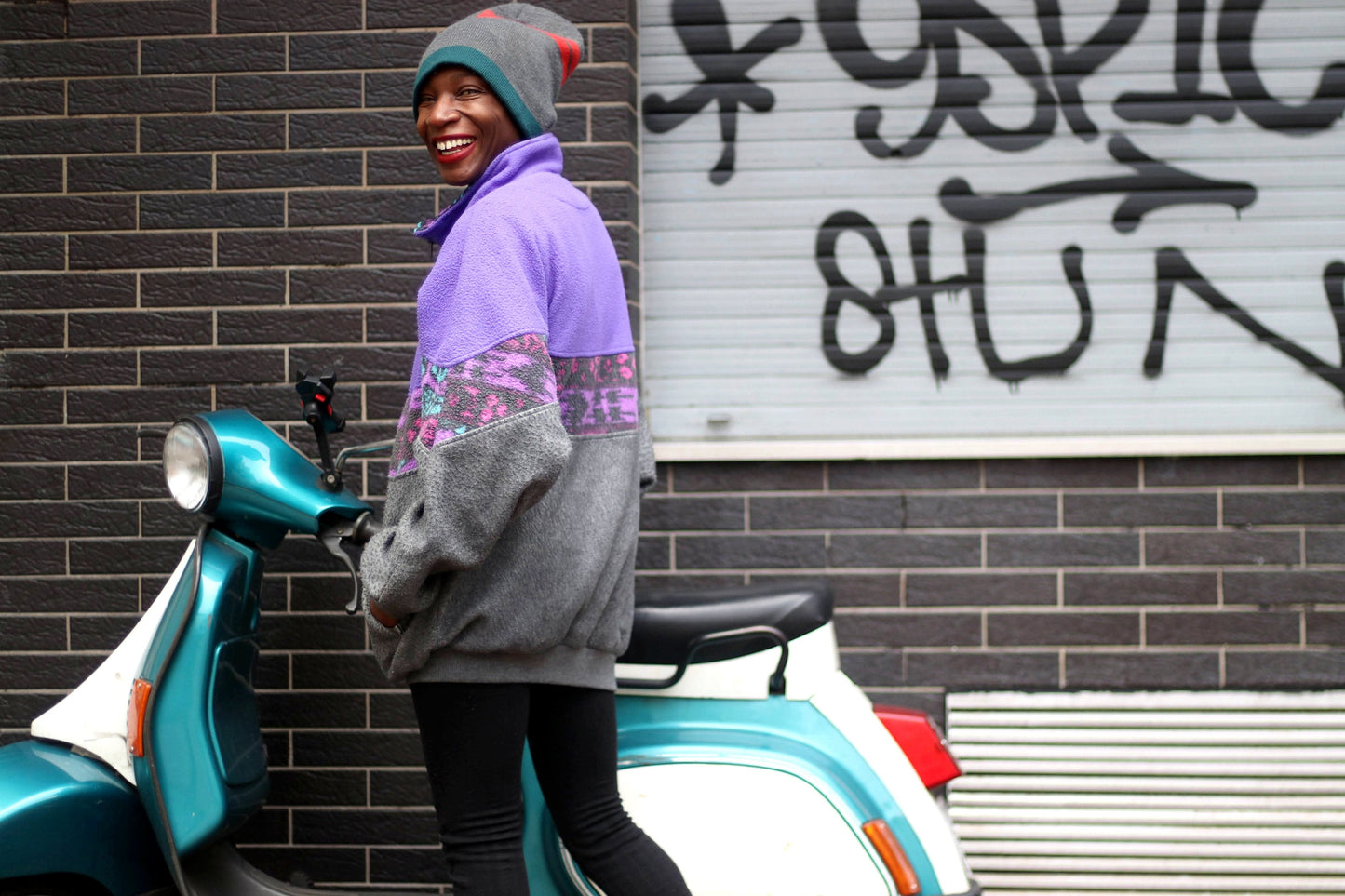 A fashionable black woman in front of a vespa packed next to a graffiti wall. She is dressed in a purple and grey zippered sweatshirt, black leggings and a striped beanie. She is smiling