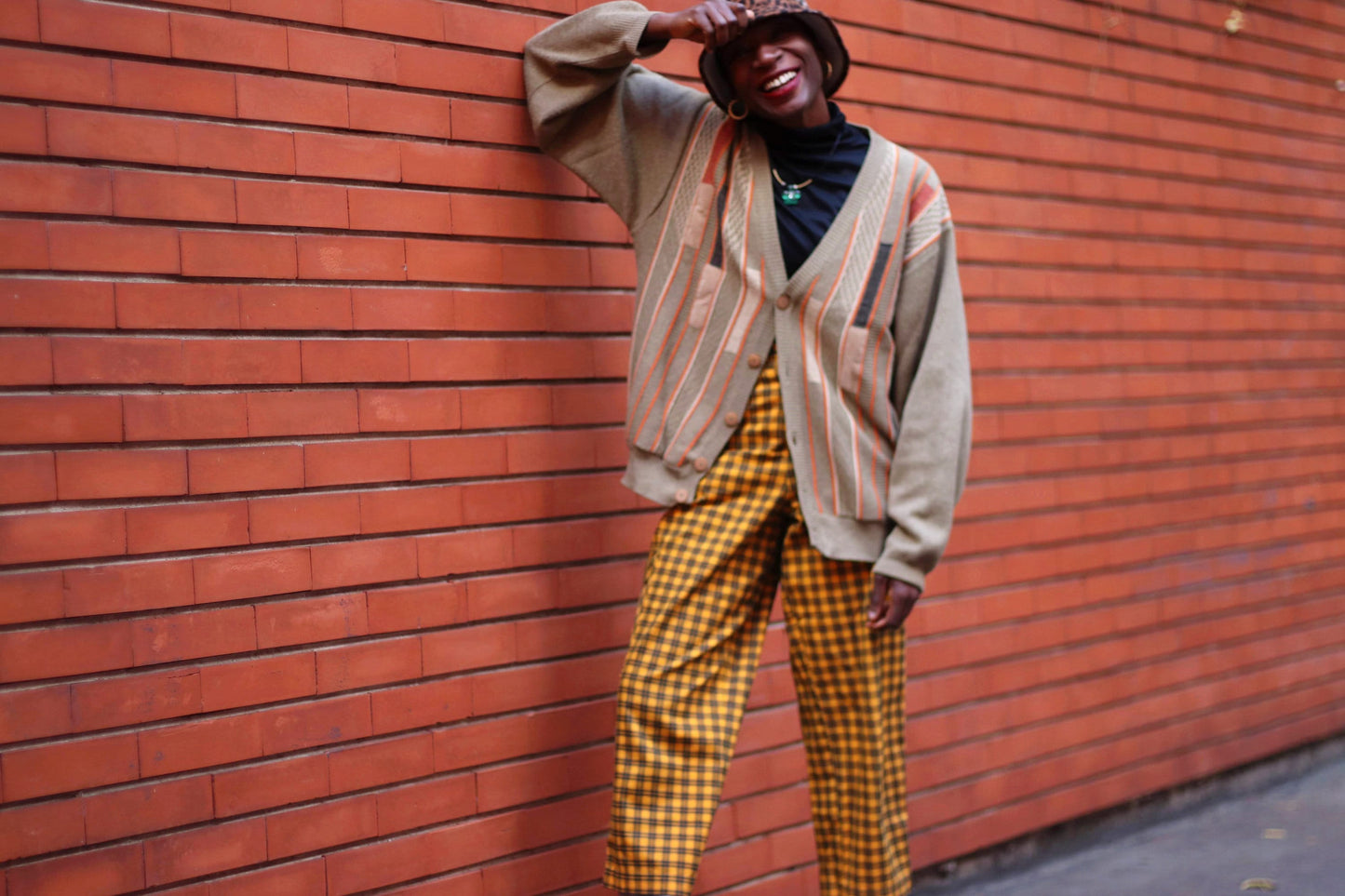 A fashionable black woman standing infront of a brick wall. One elbow rests on the wall and same hand holds her animal print hat down. She wears a black turtleneck. a brown and orange striped sweater. Her trousers are yellow with a checkered print