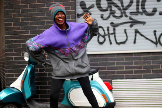 A fashionable black woman in front of a vespa packed next to a graffiti wall. She is dressed in a purple and grey zippered sweatshirt, black leggings and a striped beanie. She is smiling and waving.