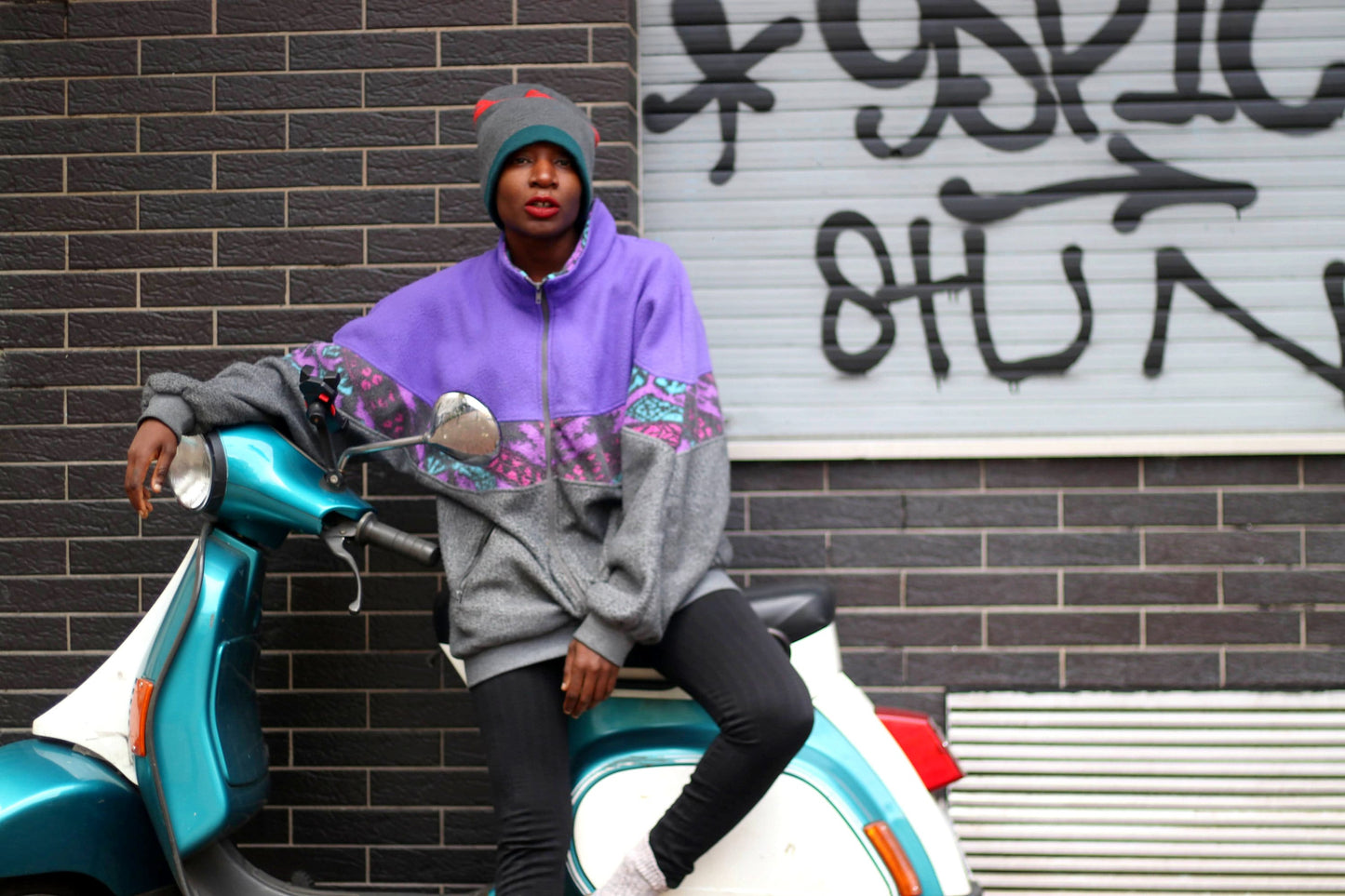 A fashionable black woman sitting sideways in front of a vespa packed next to a graffiti wall. She is dressed in a purple and grey zippered sweatshirt, black leggings and a striped beanie.