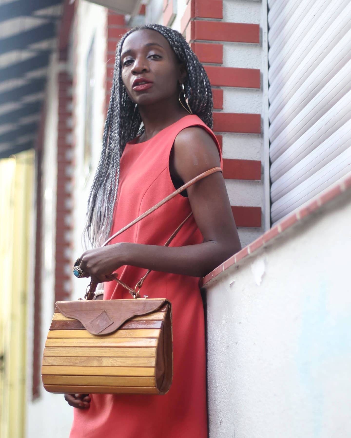 Woman in bright orange dress standing against a red-brick wall with white accents. The dress is vintage and sleeveless. She is holding a retro brown bag in one hand showcasing a feminine and timeless look.