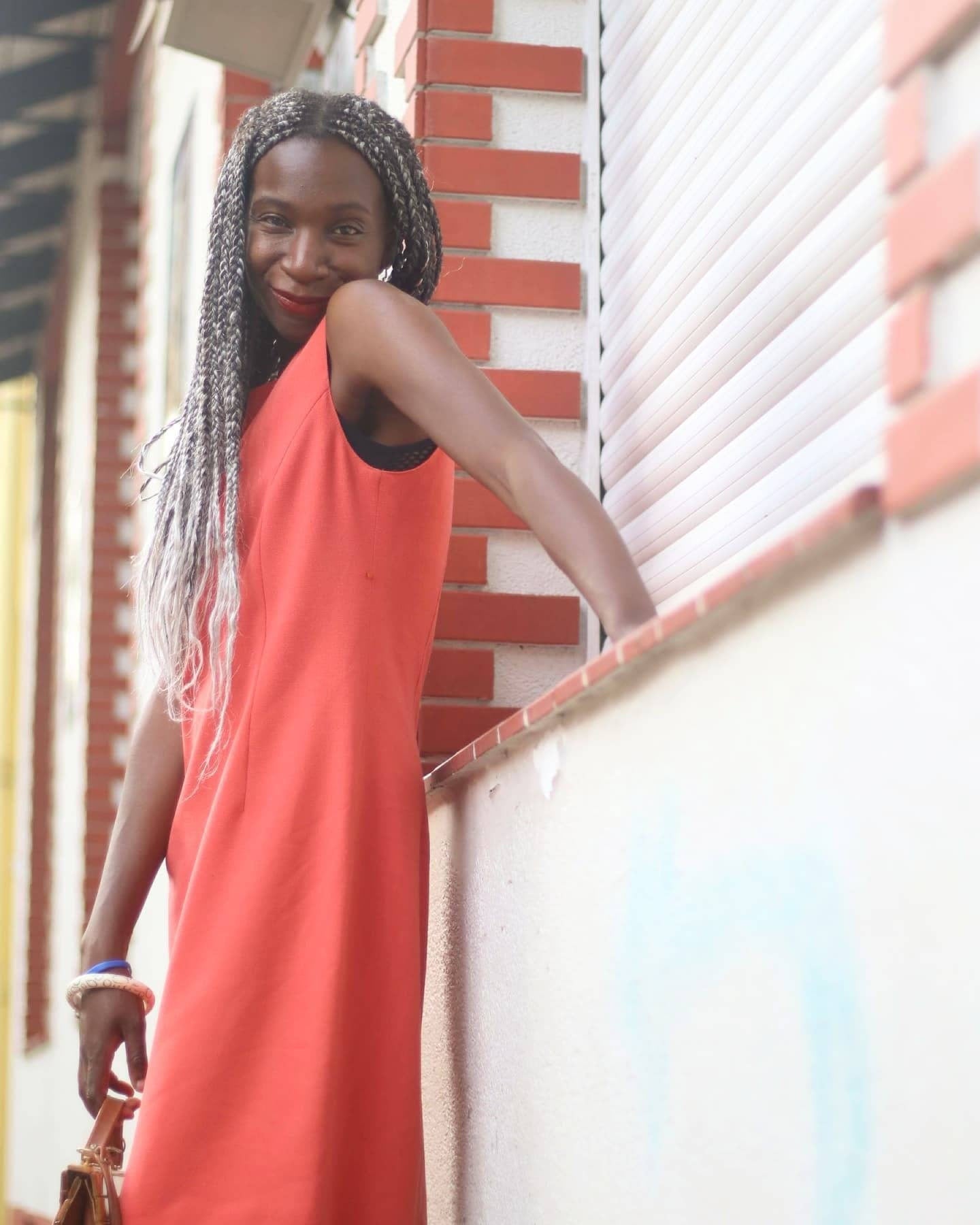 Woman in bright vintage sleeveless orange dress smiles while standing against a red-brick wall with white accents, showcasing a stylish street fashion look. She holds a brown bag