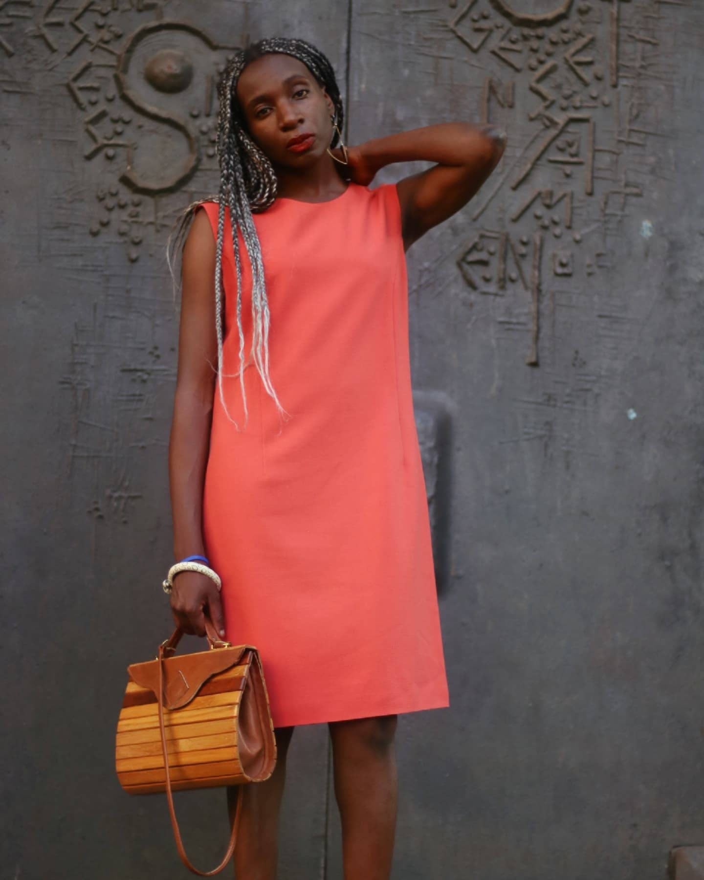Model of African heritage in a bright orange dress is photographed standing in front of an ornate grey gothic door. She has long silver-accented braids and holds a vintage wooden bag in one hand, showcasing a stylish feminine look.