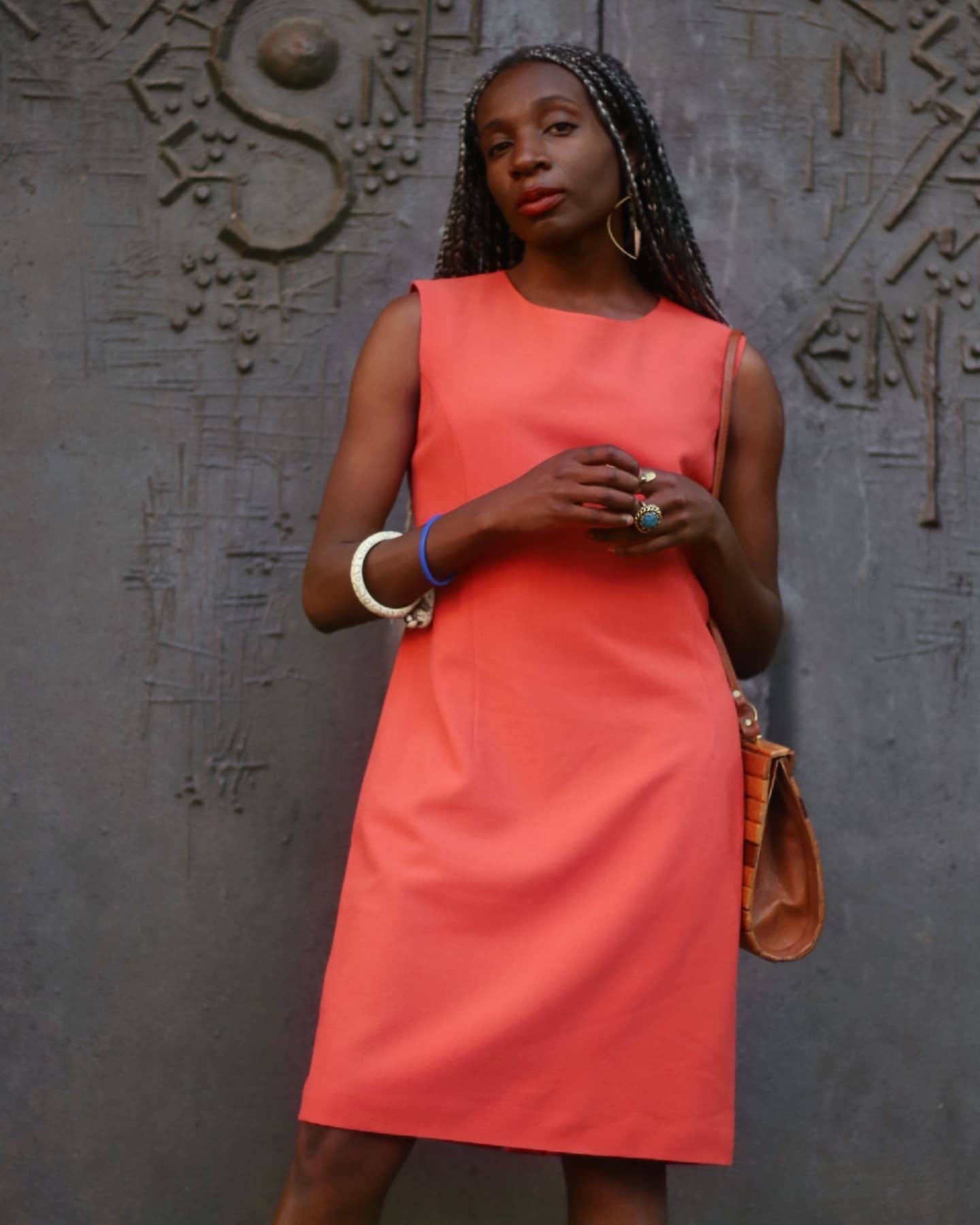 Model of African heritage in a bright orange dress is photographed standing in front of an ornate grey gothic door. She has long silver-accented braids and holds a vintage wooden bag in one hand, showcasing a stylish feminine look.