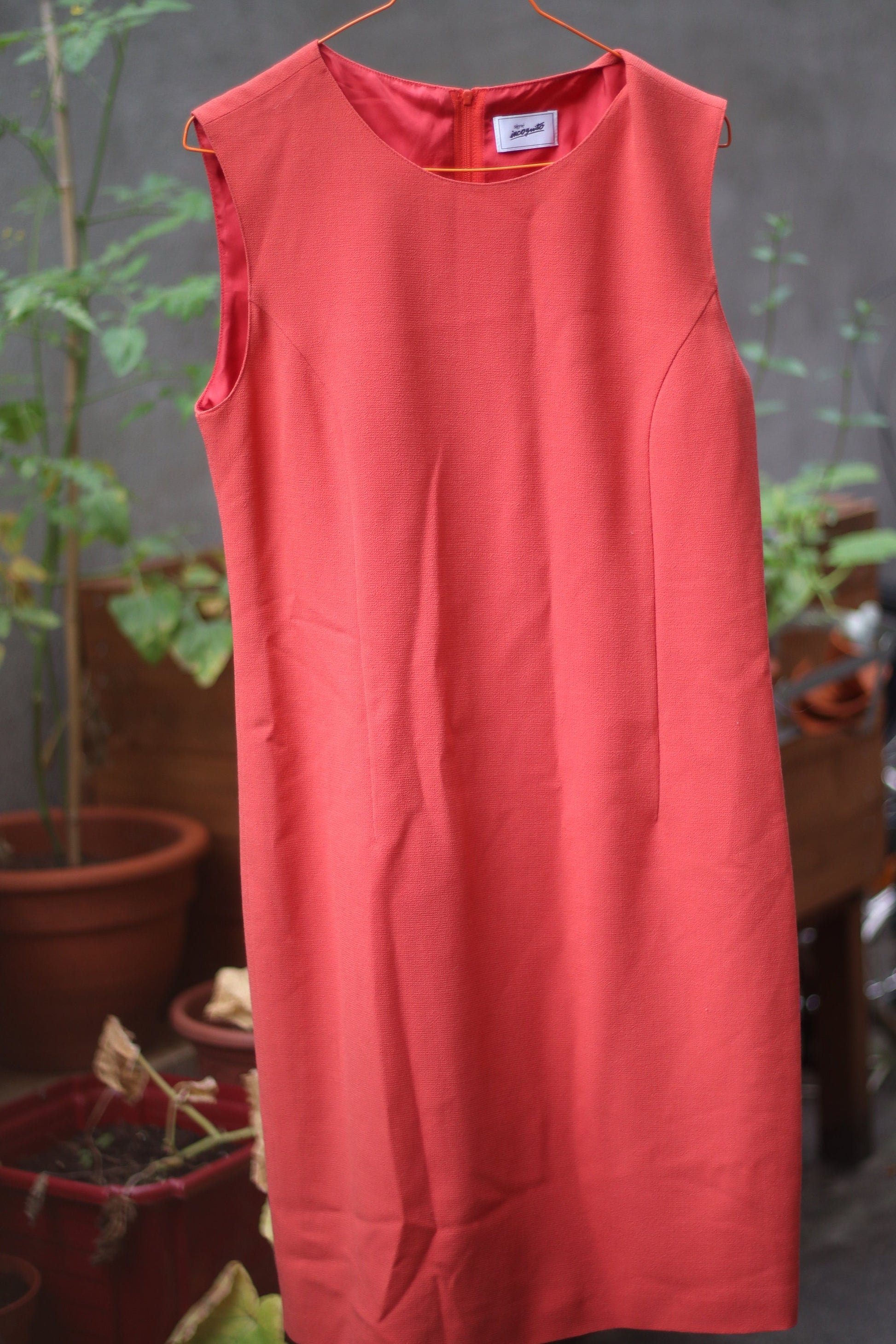 An orange sleeveless vintage dress by the brand Signe incognito is photographed outdoors on a red hanger against a leafy background.