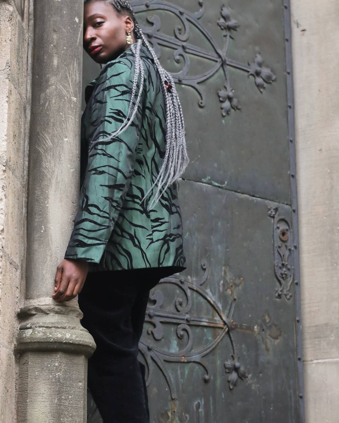 Model of African heritage wearing a vintage green and black zebra-print blazer, tilting their head while posing against an ornate dark green wrought-iron door.