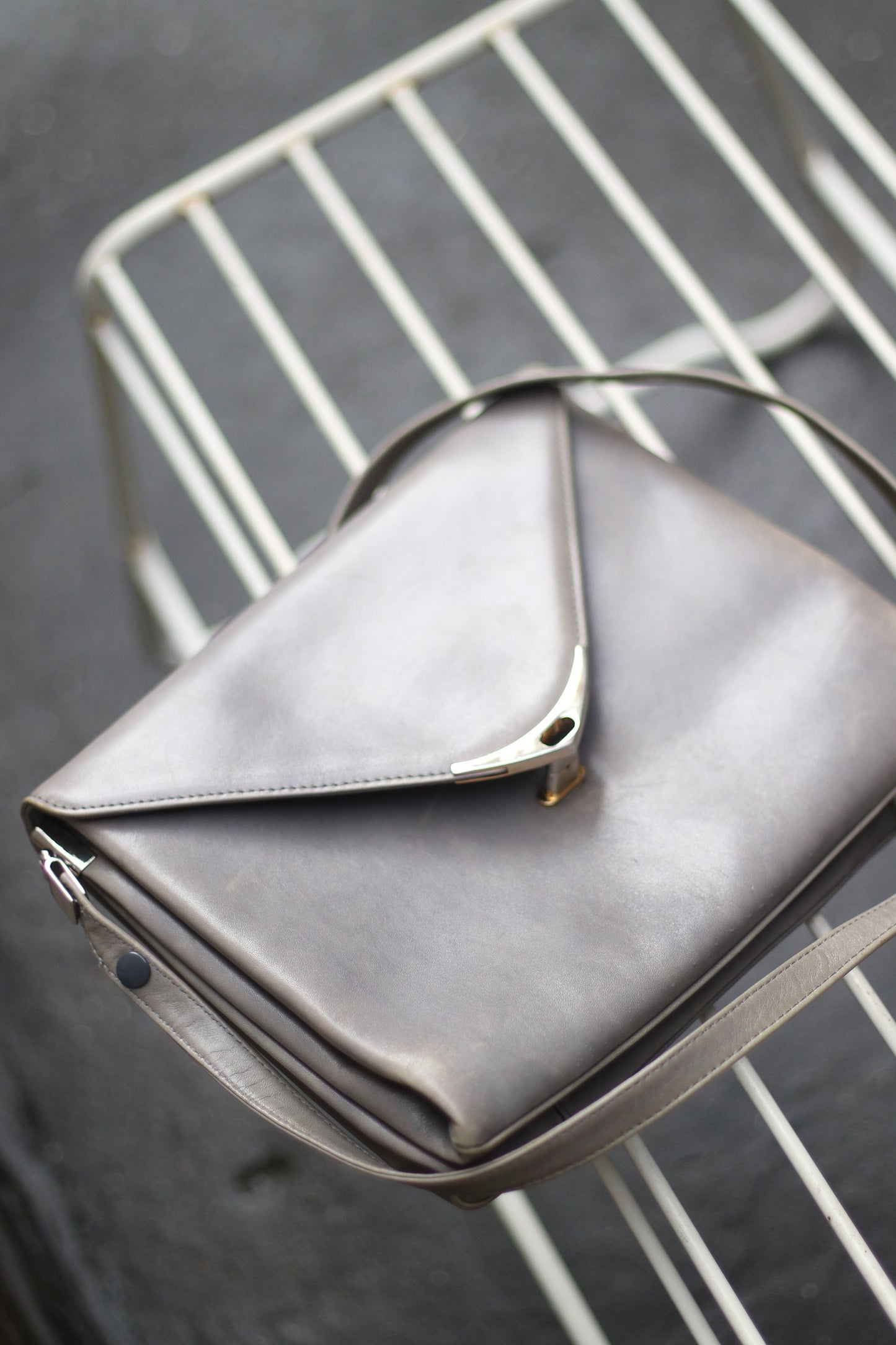 A grey compact bag from the 70s photographed against on a white lined stool. The minimalist leather bag for women is by the german brand Nierhaus. Small in size, the bag can be carried crossbody on on the shoulder.