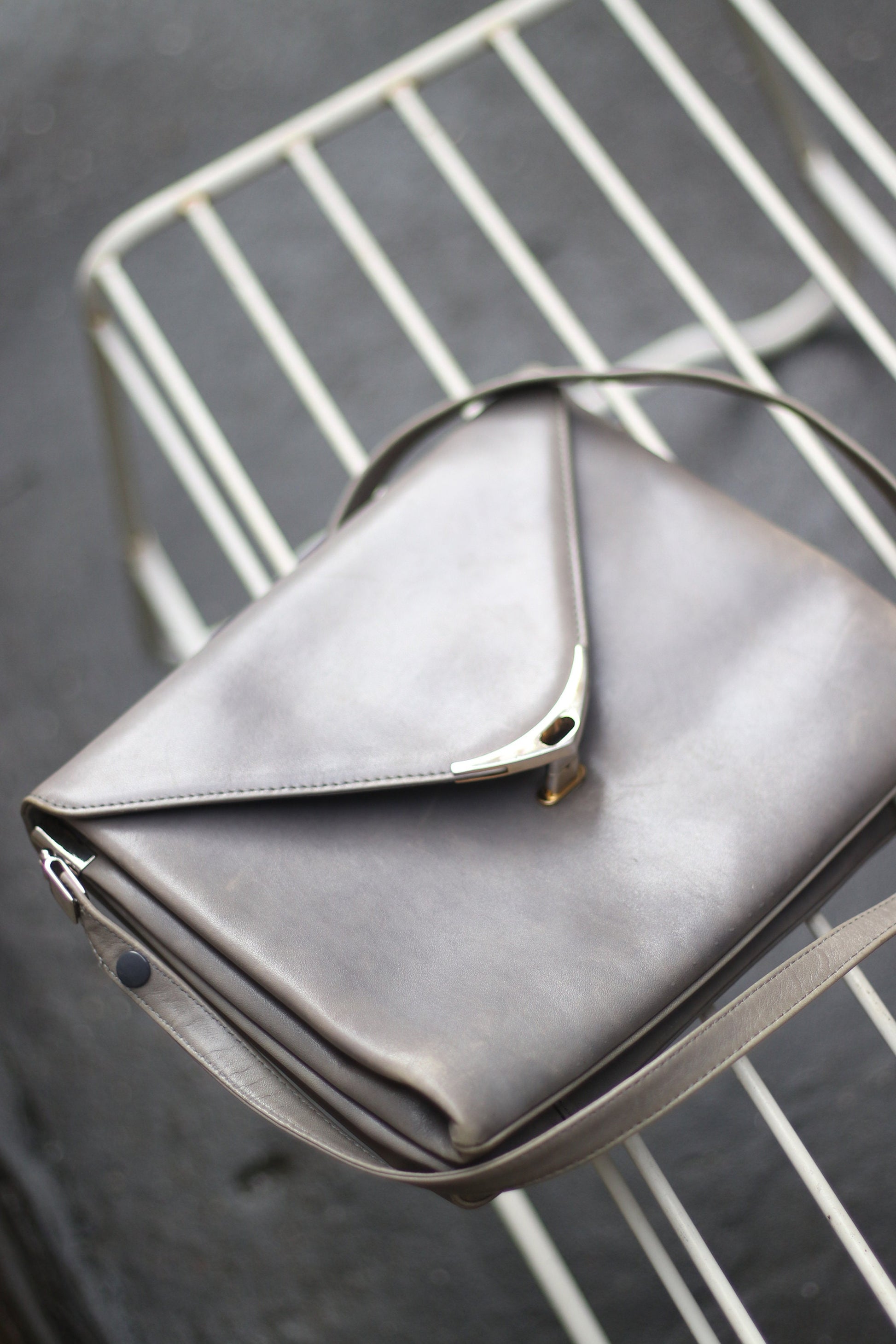 A grey compact bag from the 70s photographed against on a white lined stool. The minimalist leather bag for women is by the german brand Nierhaus. Small in size, the bag can be carried crossbody on on the shoulder.