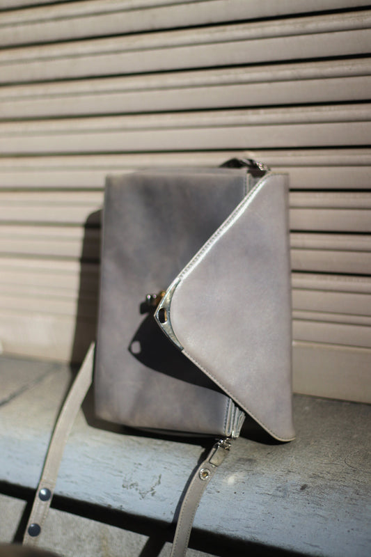 A grey compact bag from the 70s photographed against a cream lined urban background. The minimalist leather bag for women is by the german brand Nierhaus. Small in size, the bag can be carried crossbody on on the shoulder.