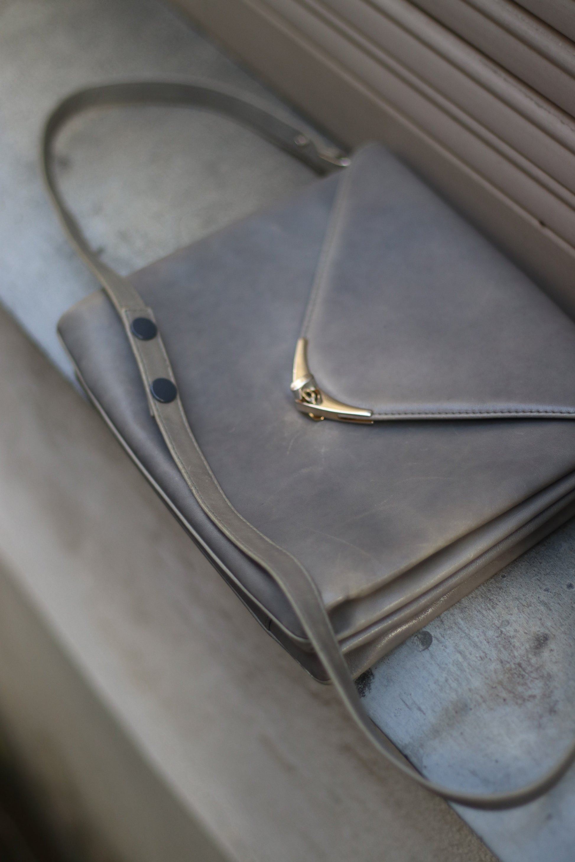 A grey compact bag from the 70s photographed lying flat a grey slate stone. The minimalist leather bag for women is by the german brand Nierhaus. Small in size, the bag has adjustable straps.