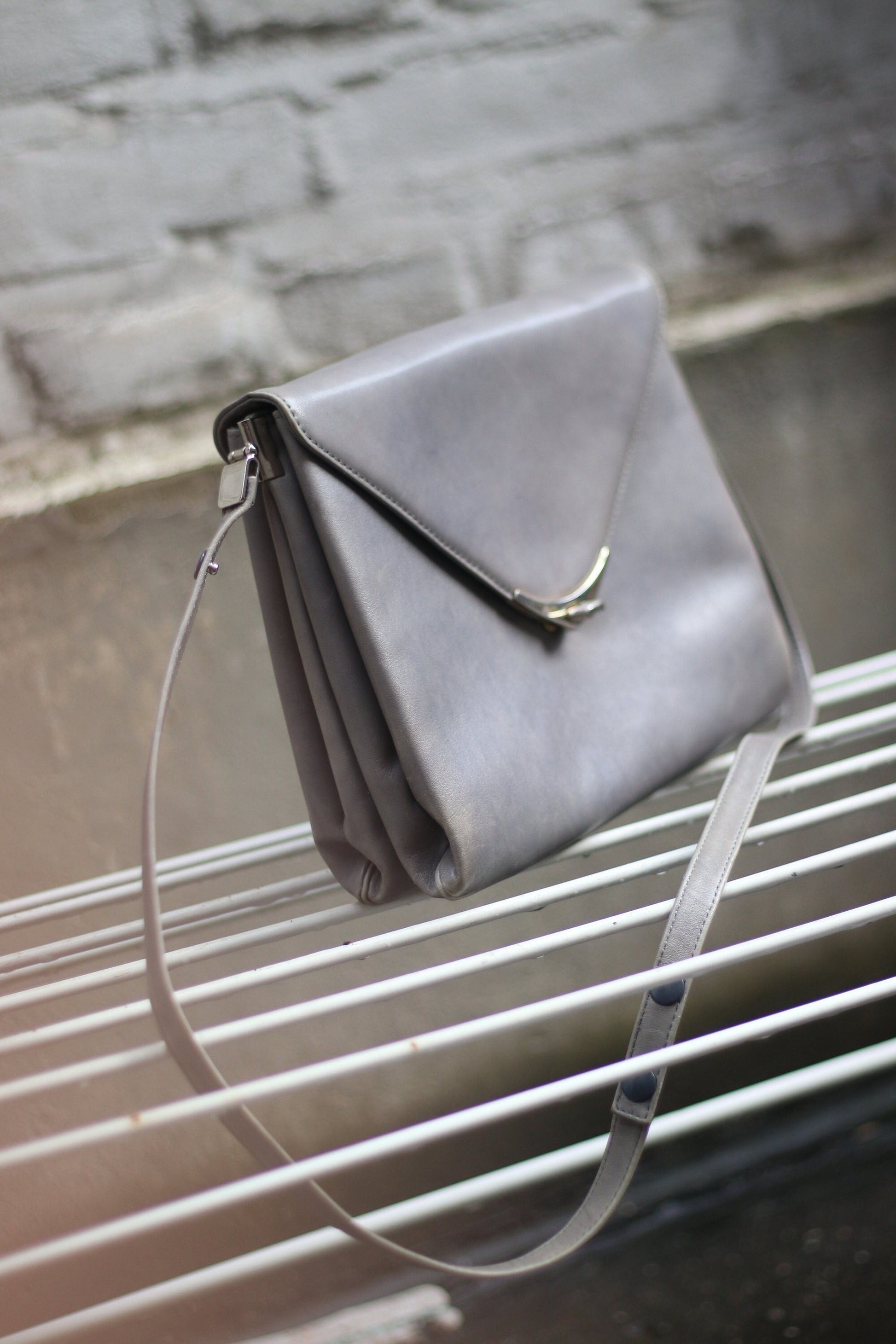 A grey compact bag from the 70s photographed against a white brick wall on a white lined stool. . The minimalist leather bag for women is by the german brand Nierhaus. Small in size, the bag can be carried crossbody on on the shoulder.