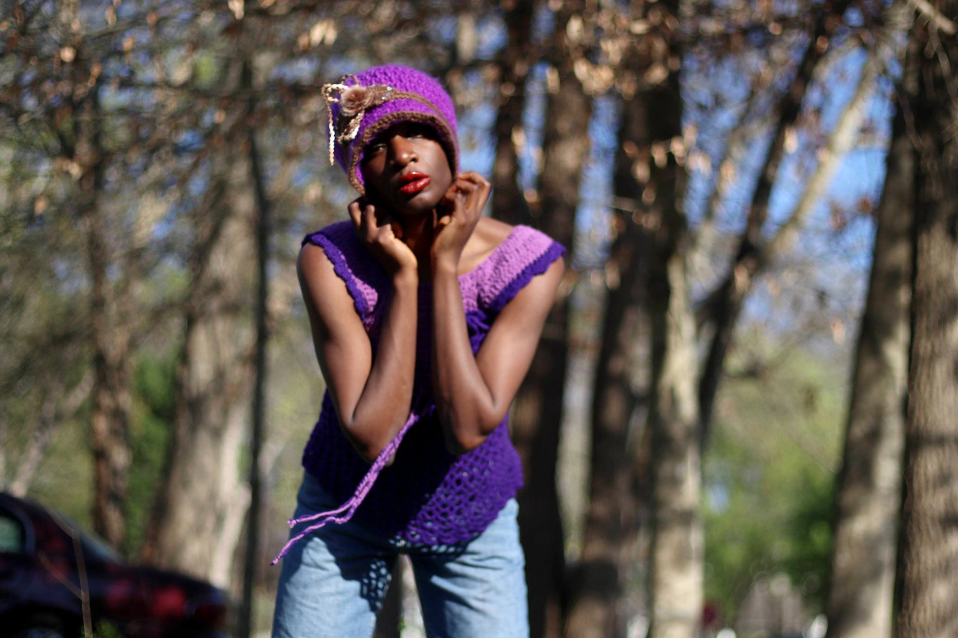 Model of African heritage in a vintage handmade purple crochet top and jeans posing against a background with green trees, outdoor setting. She is wearing a matching purple hat