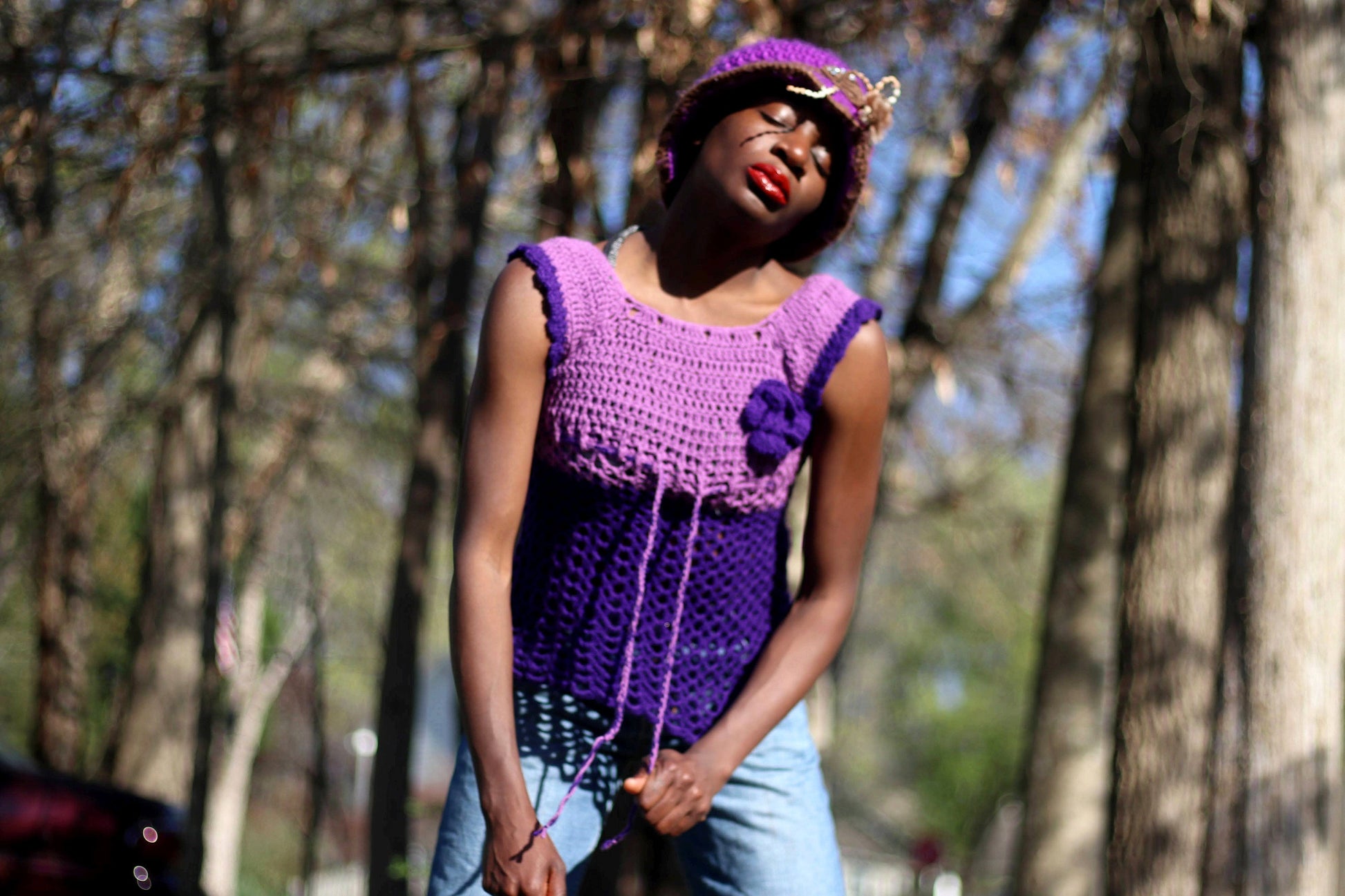 Model of African heritage in a vintage handmade purple crochet top and jeans posing against a background with green trees, outdoor setting. She is wearing a matching purple hat and eyes are closed