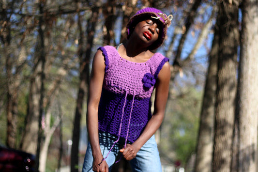 Model of African heritage in a vintage handmade purple crochet top and jeans posing against a background with green trees, outdoor setting. She is wearing a matching purple hat and eyes are closed