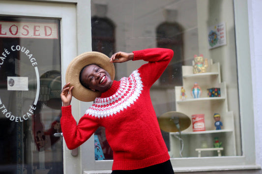 Black female model in a vintage Scandinavian red sweater with white patterns and wide-brimmed hat smiling and posing happily in front of a shop window with a closed sign, enjoying a fun moment outdoors.