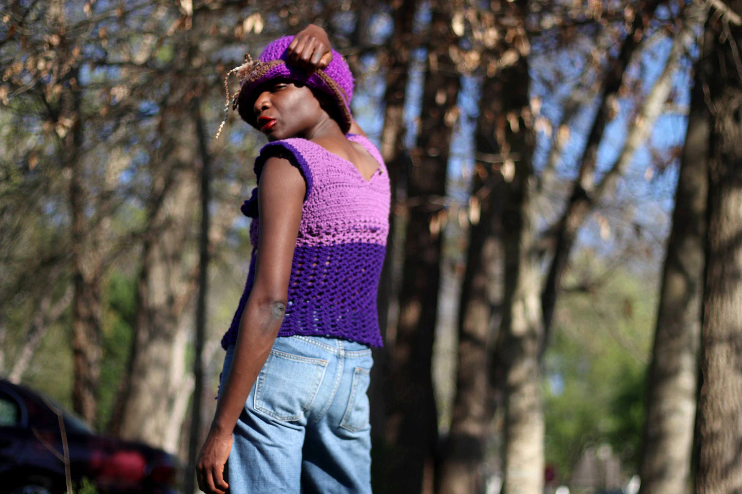 Model of African heritage in a vintage handmade purple crochet top and jeans posing against a background with green trees, outdoor setting. She is wearing a matching purple hat and eyes are closed