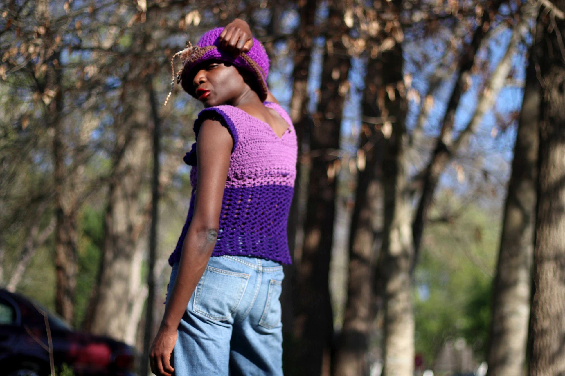 Model of African heritage in a vintage handmade purple crochet top and jeans posing against a background with green trees, outdoor setting. She is wearing a matching purple hat and eyes are closed