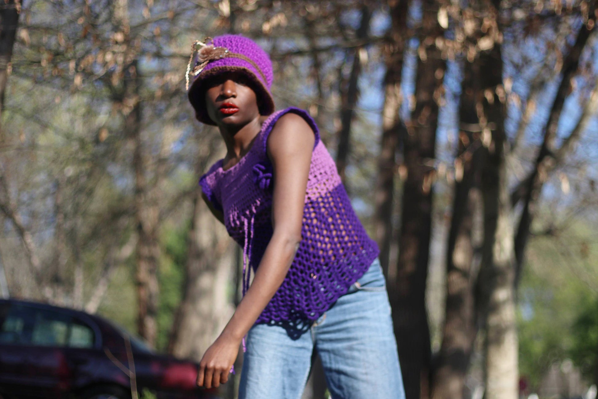 Model of African heritage in a vintage handmade purple crochet top and jeans posing against a background with green trees, outdoor setting. She is wearing a matching purple hat