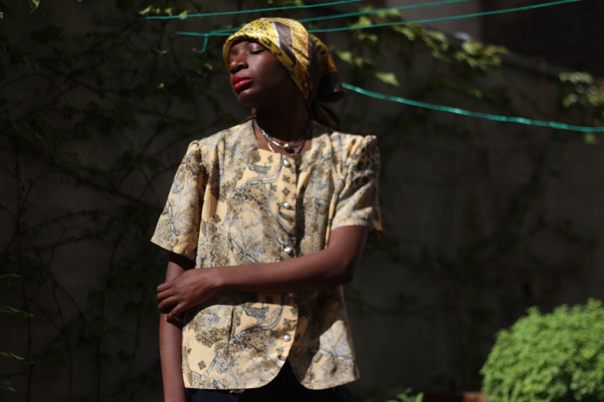 A Black Model in an 80s Paisley Print Brown blouse with short sleeves. She has a loosely tied headscarf on and photographed against a walled background with plants .