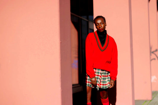 Model of African Heritage in a  vintage v neck collegiate chic red sweater and a mini plaid skirt standing against a pink wall with shadows.