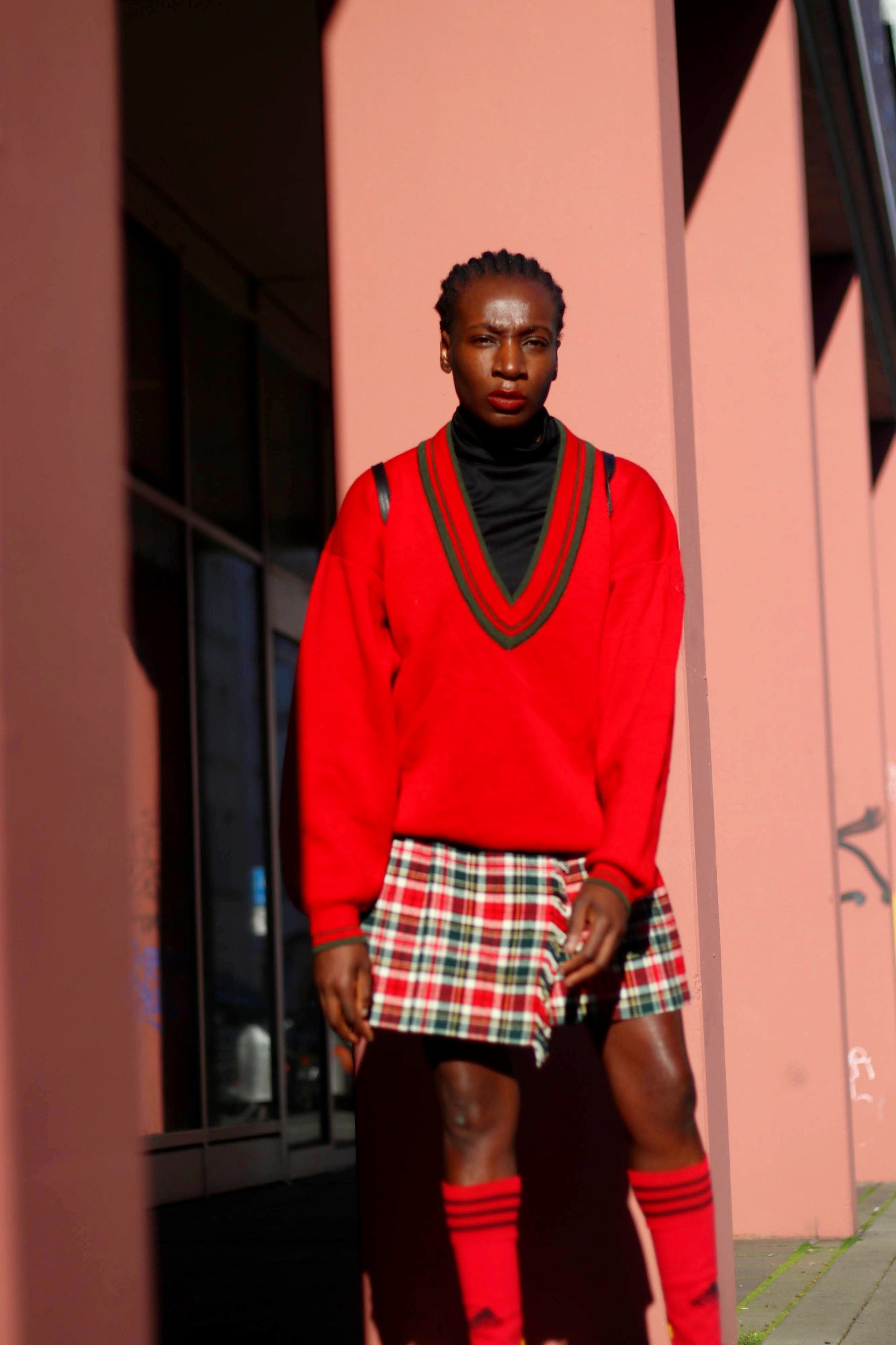 Model of African Heritage in a  vintage v neck collegiate chic red sweater and a mini plaid skirt standing against a pink wall with shadows. She also swears calf-length  red socks