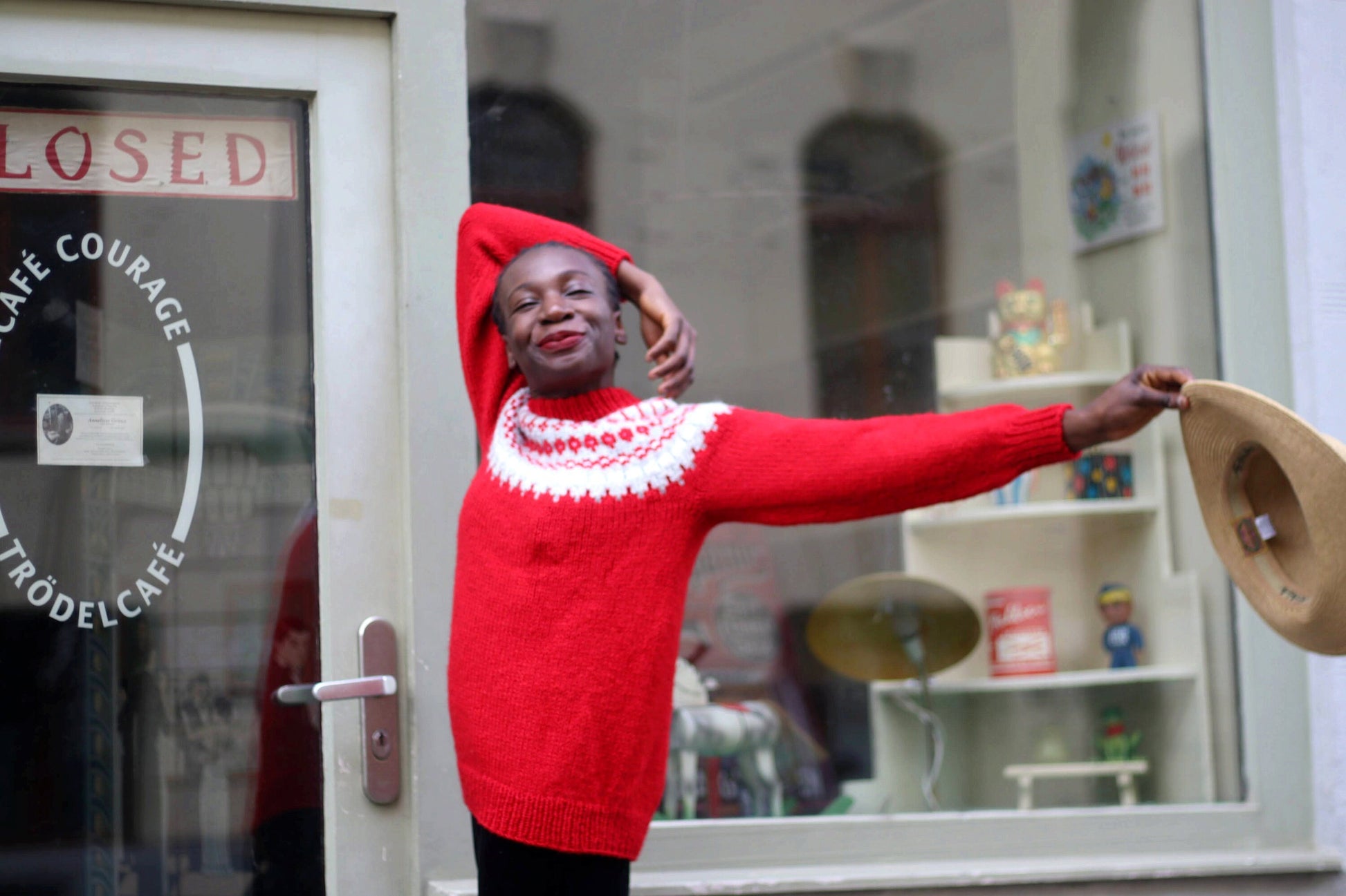 Female model of African heritage in a vintage red Scandinavian long sleeved jumper with white motifs by the collar. She is holding a wide-brimmed hat smiling and posing happily in front of a shop window with a closed sign.