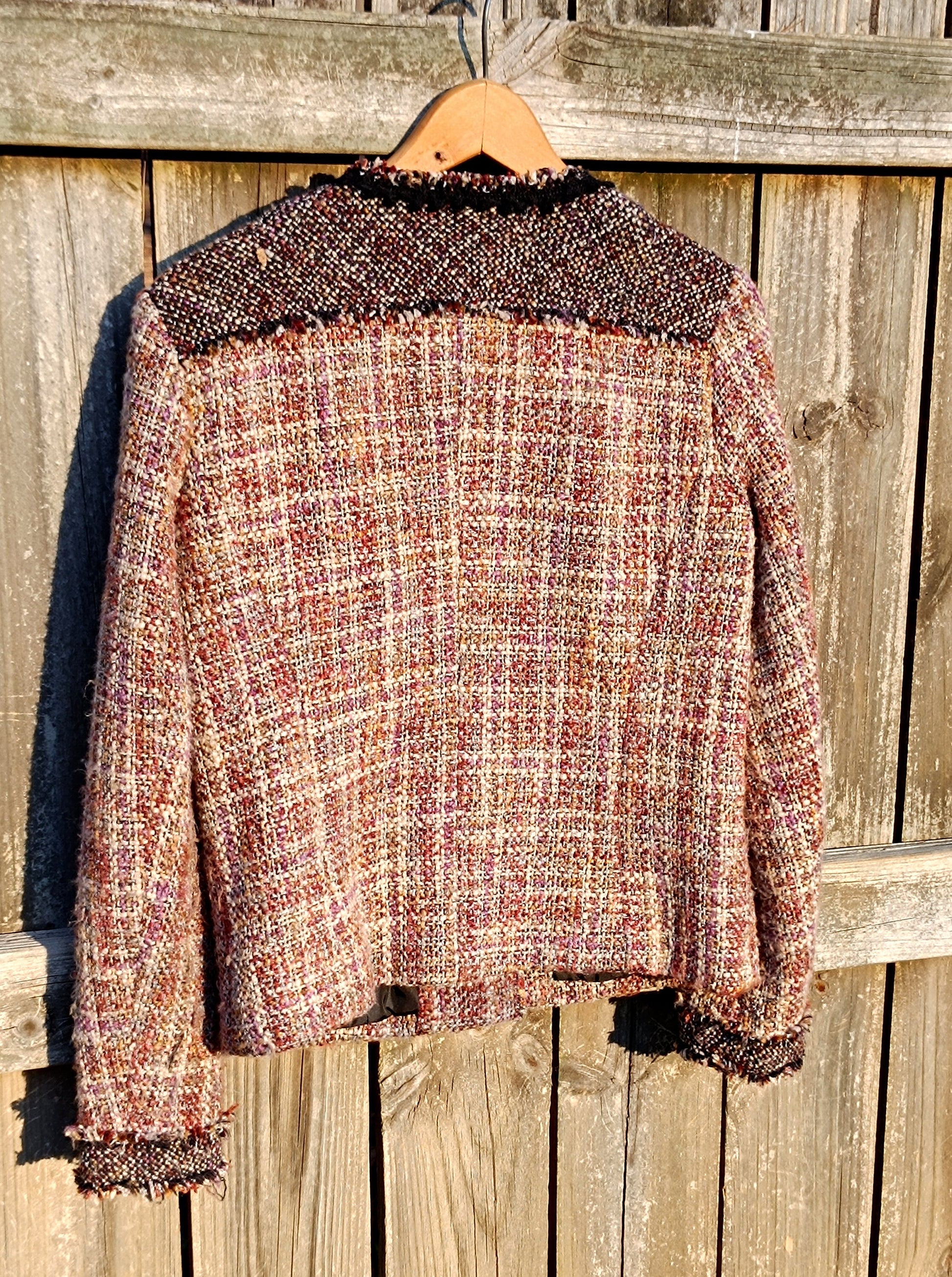 The back view of a vintage textured tweed blazer from the 90s in shades of pink, cream, and black hanging against a rustic wooden fence.