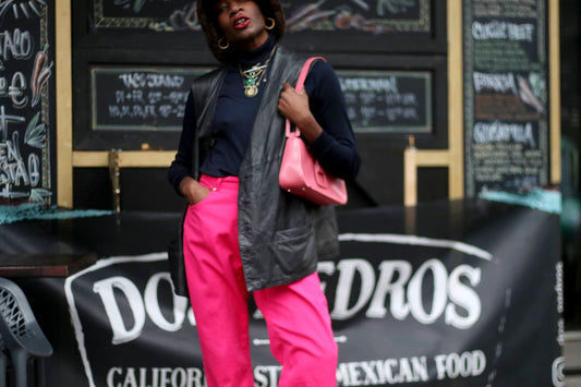 model poses in a pair of pink Escada Sport high-waisted trousers, styled with a black turtle neck, a leather vest and a bold pink handbag Infront of a restaurant with a menu board in the background .