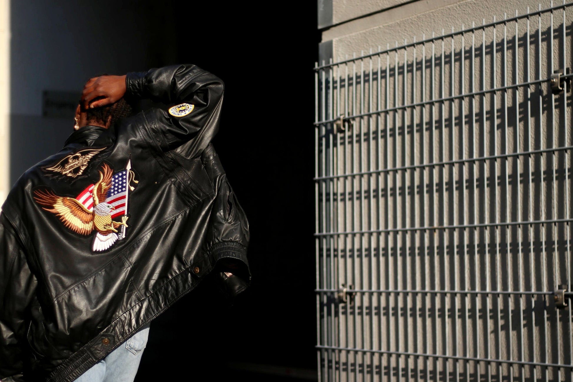 Person of African Heritage wearing a black vintage biker leather jacket with an embroidered eagle and American flag design, standing beside a metal fence. Street style fashion with casual urban backdrop