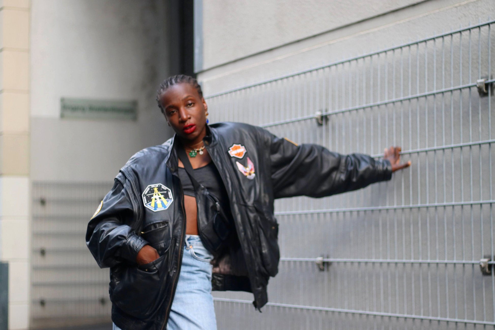 Woman of African heritage in a Vintage Black Leather Jacket and blue Jeans Leaning Against Metal Fence, Jacket has Patchwork Details Street Fashion Photography.