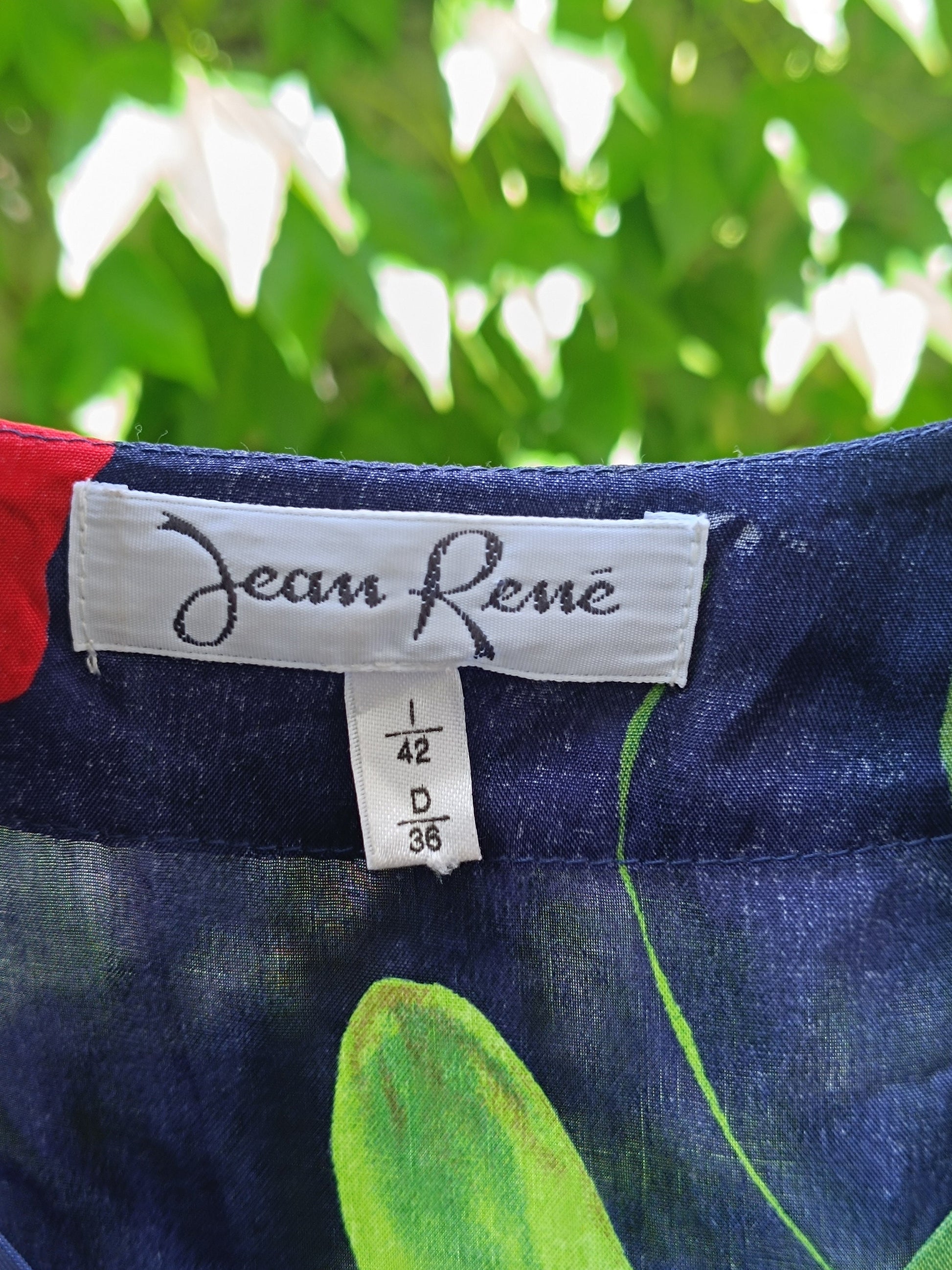 Front view of the brand tag of a vintage navy floral blouse with bold hibiscus print. The tag reads &quot; Jean Rene&quot;  also visible is the size tag