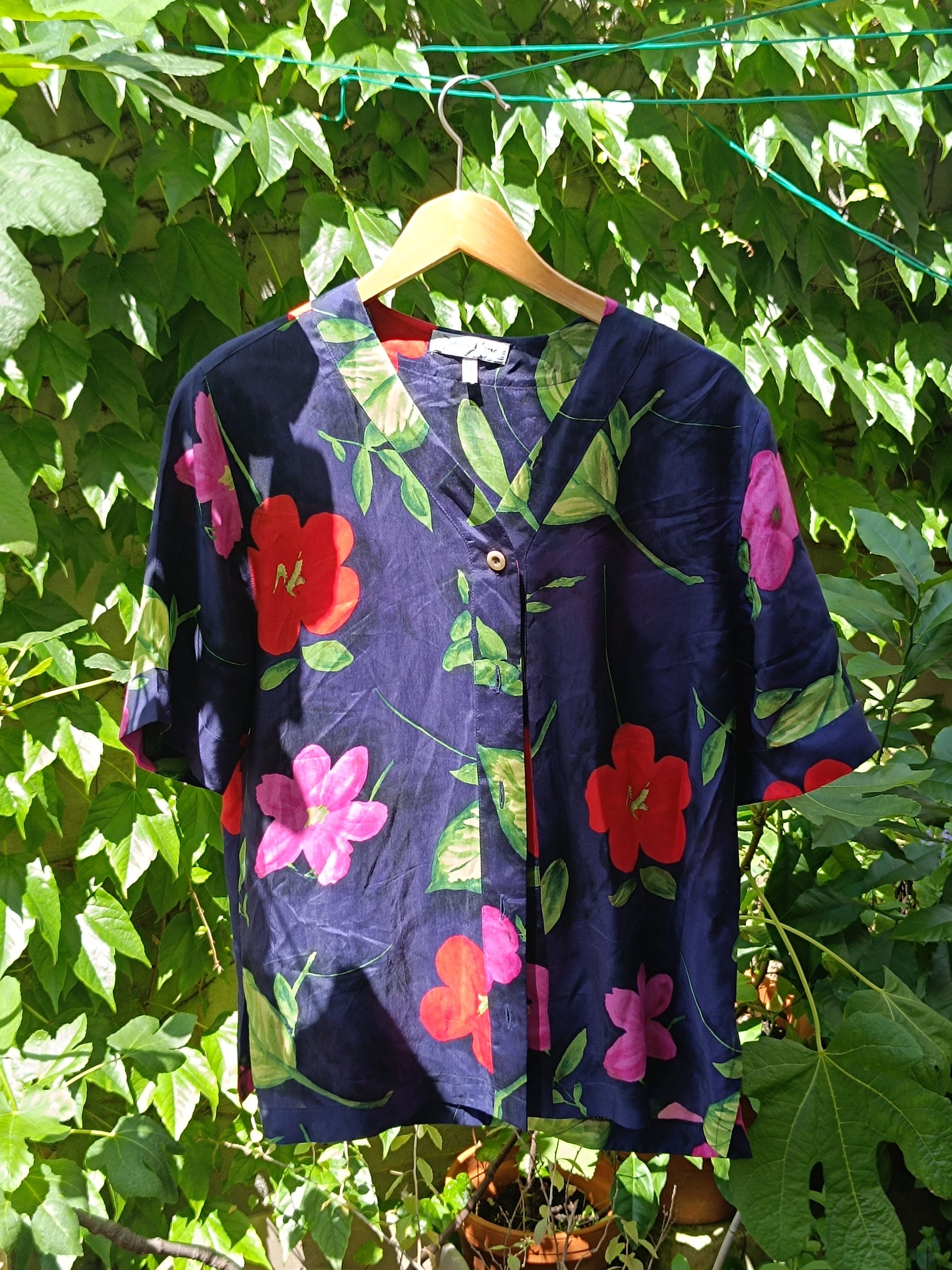 A 90s  navy floral blouse with bold hibiscus print photographed on a red hanger Infront of a leafy background . The image focuses on the v-neck silhouette of the button-up