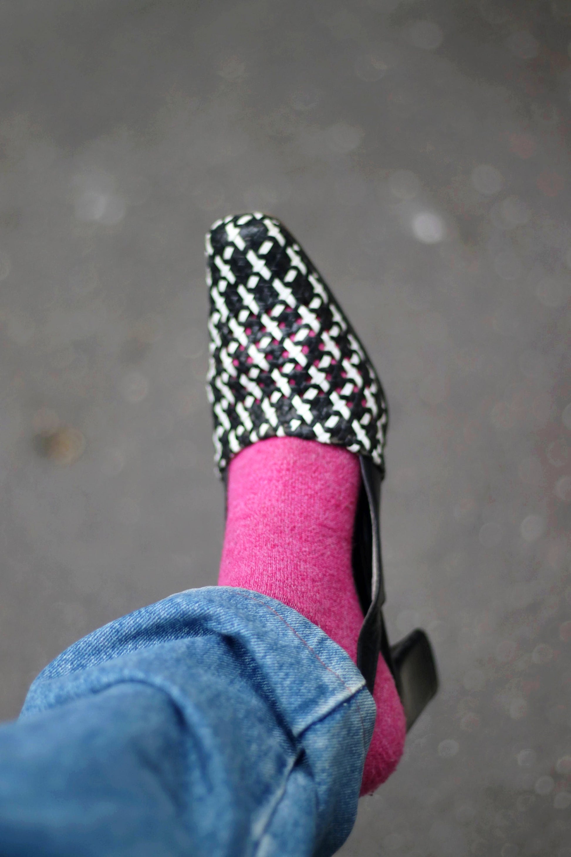 A single vintage black and white woven leather slingback shoe is shown on a foot with pink shoes. The shoe features a pointed toe, a low block heel, and an intricate woven pattern.The background is a blurry gray asphalt surface.