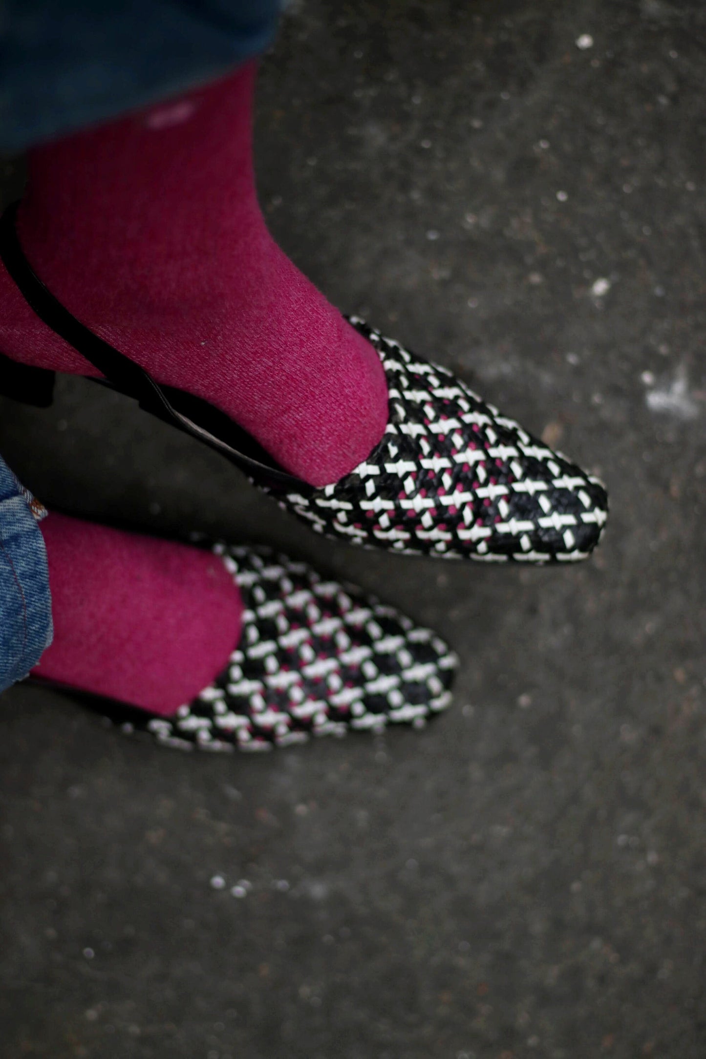Close-up, top-down view of two vintage black and white woven leather heels, showcasing the intricate weave pattern.  The pointed toes and low block heels are visible. pink socks are visible inside the shoes. The background is a textured gray surface.