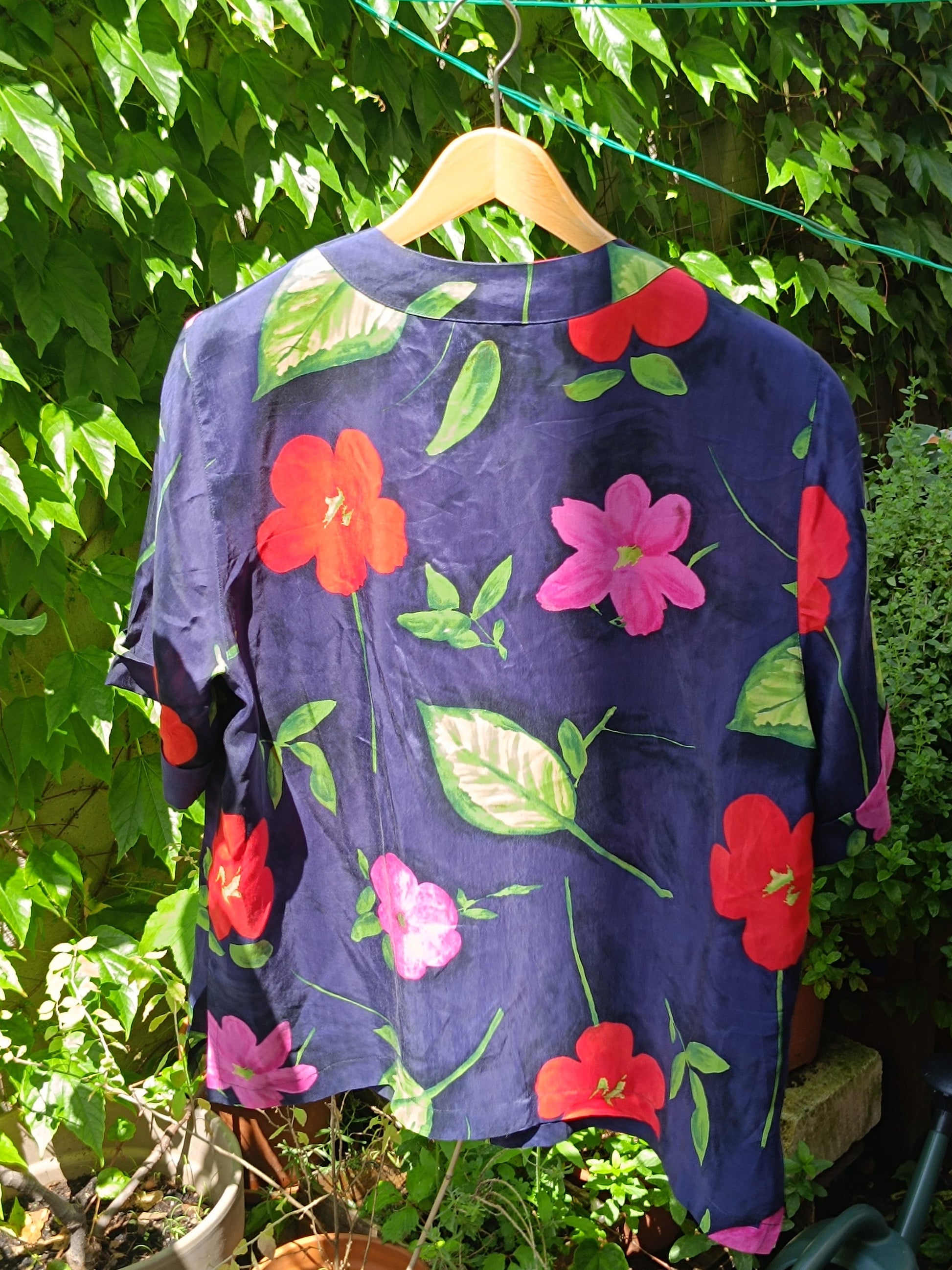 The back view of a vintage navy floral blouse with bold hibiscus print photographed on a red hanger Infront of a leafy background in a garden. The image highlights the  summer ready patterns