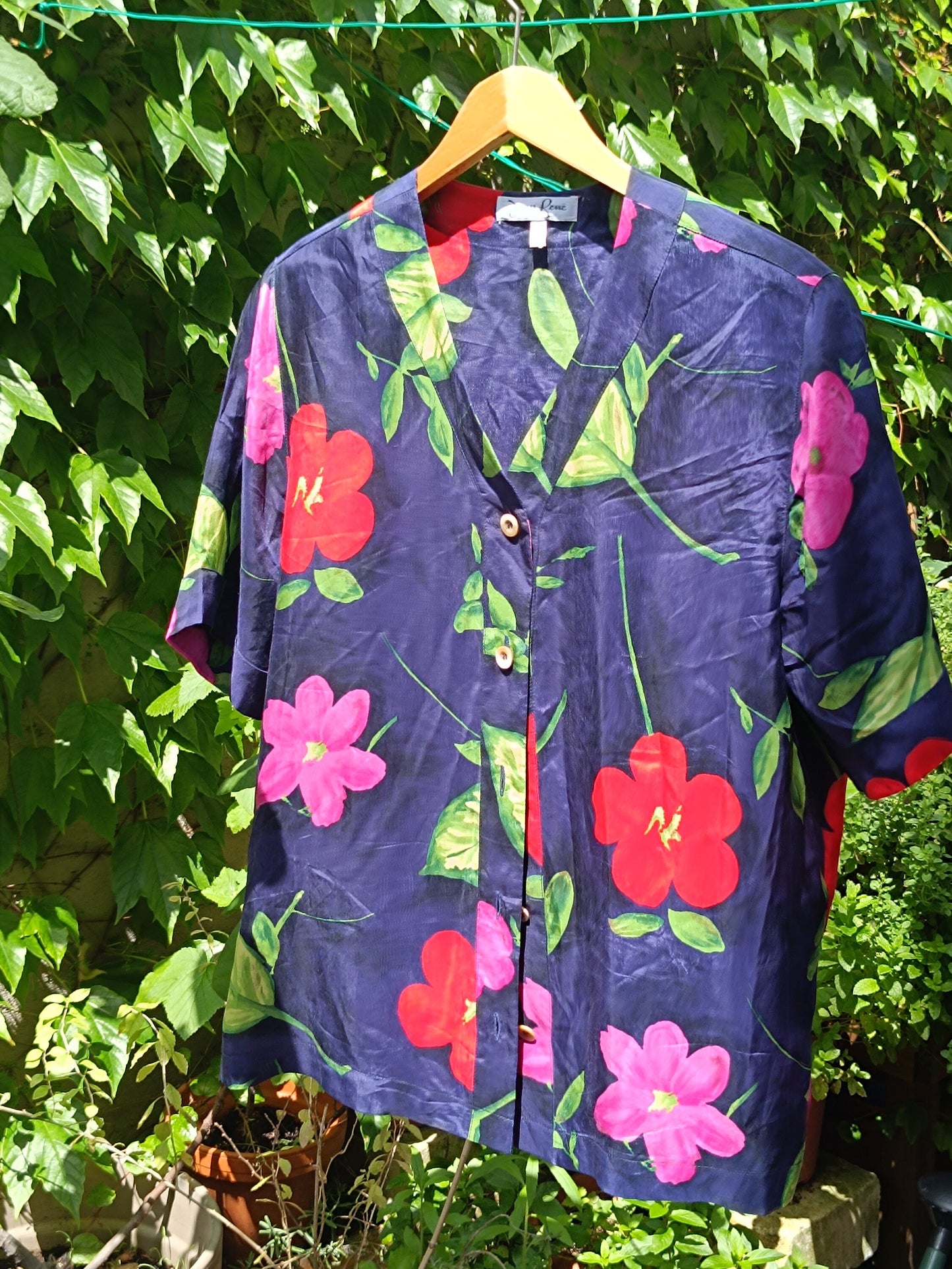 A vintage navy floral blouse with bold hibiscus print photographed on a red hanger Infront of a leafy background in a garden. The image focuses on the statement  print of the 90s summer shoet-sleeved design