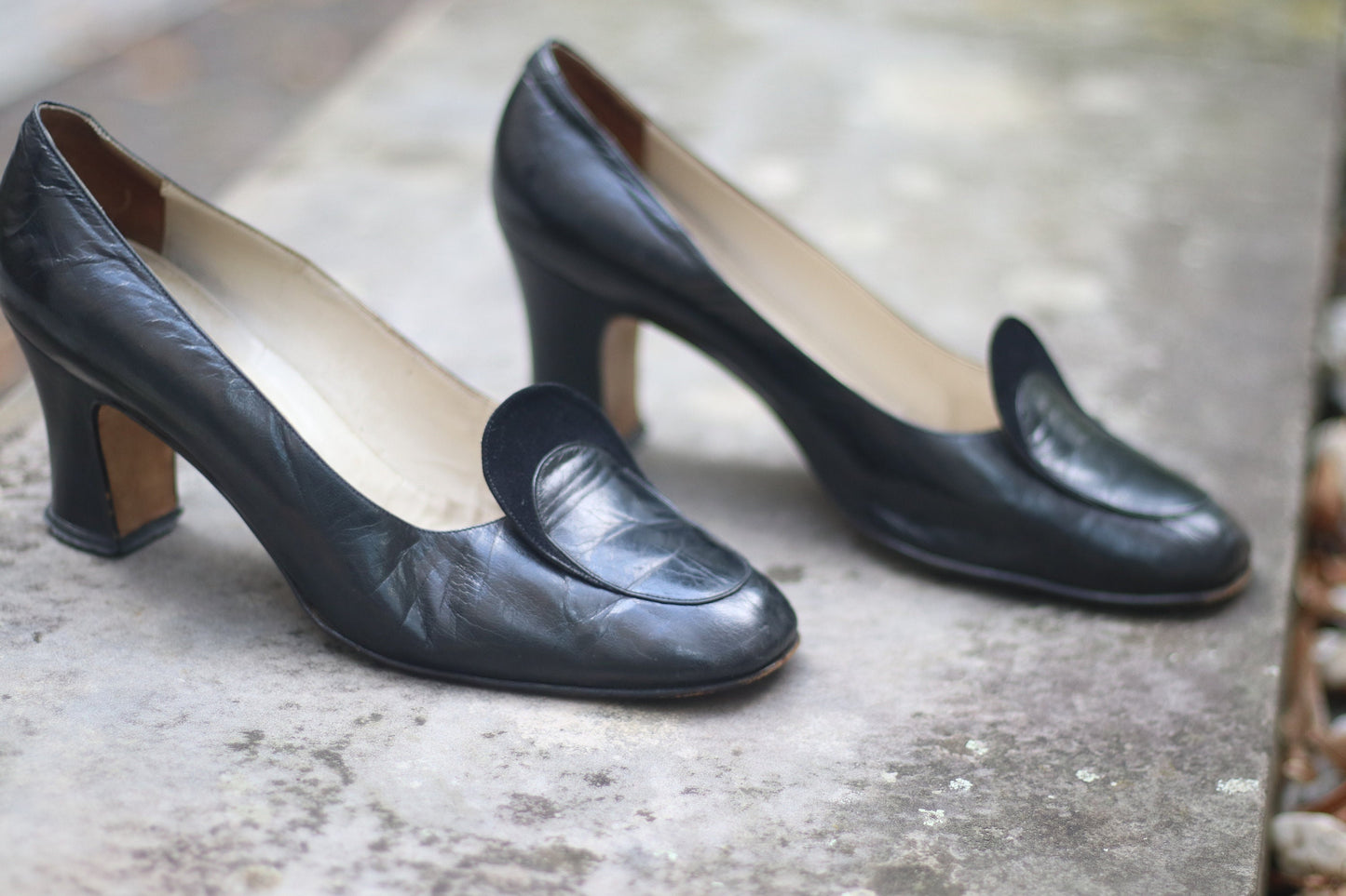 Vintage black leather high-heeled shoes with a rounded toe and mid heel, showcased on a concrete surface. Perfect for retro fashion enthusiasts and classic footwear collectors.