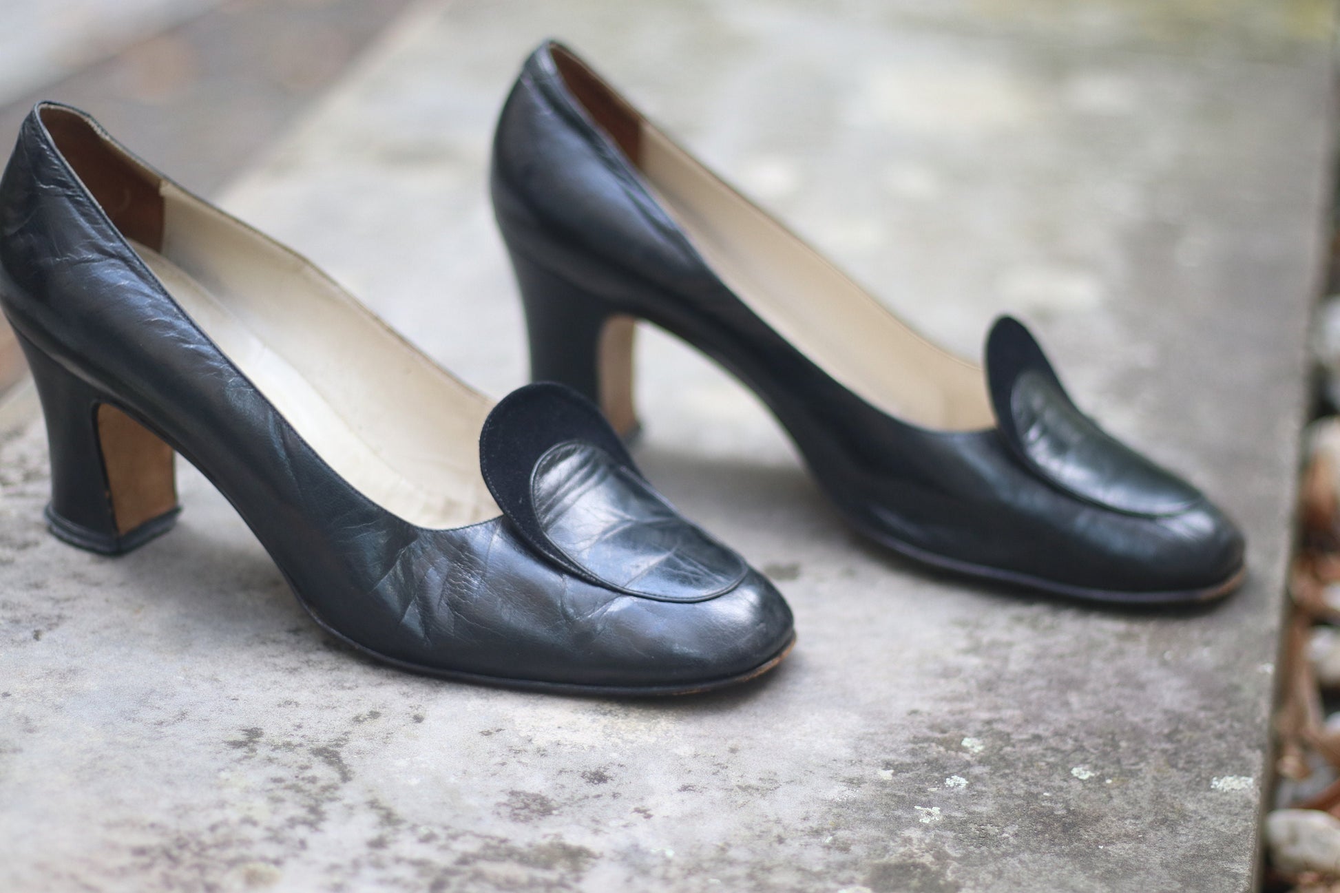 Vintage black leather high-heeled shoes with a rounded toe and mid heel, showcased on a concrete surface. Perfect for retro fashion enthusiasts and classic footwear collectors.
