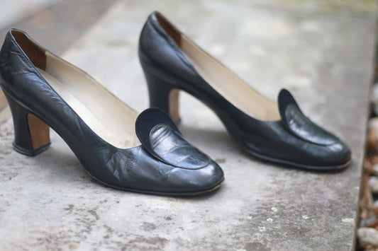 Vintage black leather high-heeled shoes with a rounded toe and mid heel, showcased on a concrete surface. Perfect for retro fashion enthusiasts and classic footwear collectors.