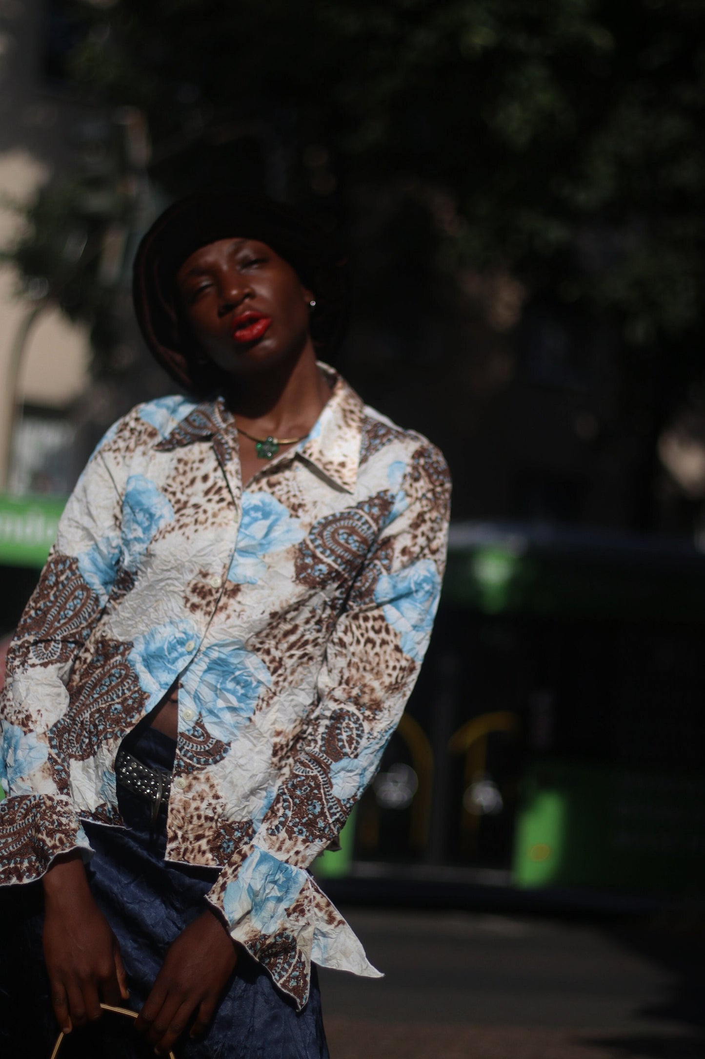 Model of African Heritage in a vintage y2k floral print shirt with butterfly sleeves and dark skirt, carrying a beige shoulder bag standing in sunlight.