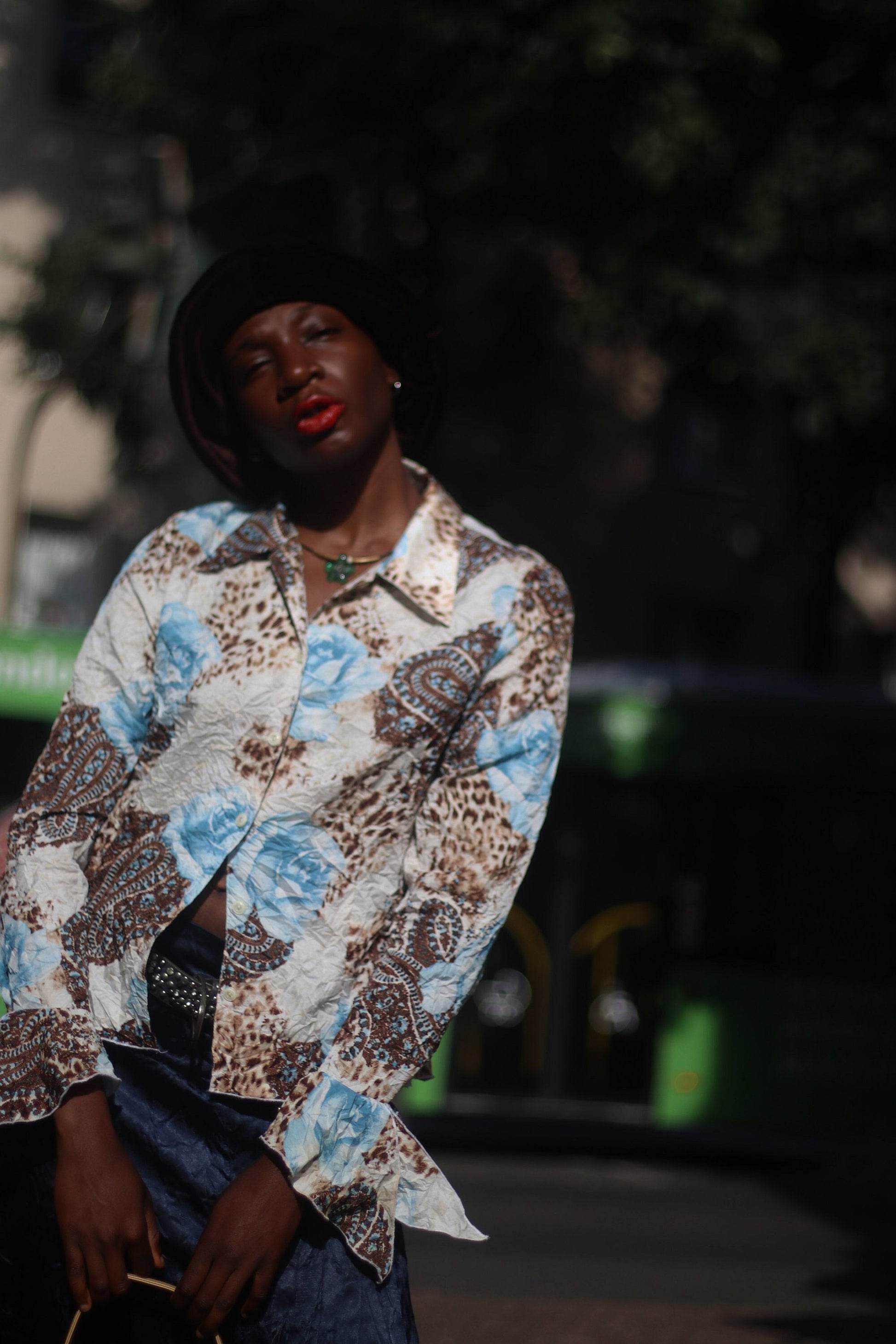 Model of African Heritage in a vintage y2k floral print shirt with butterfly sleeves and dark skirt, carrying a beige shoulder bag standing in sunlight.